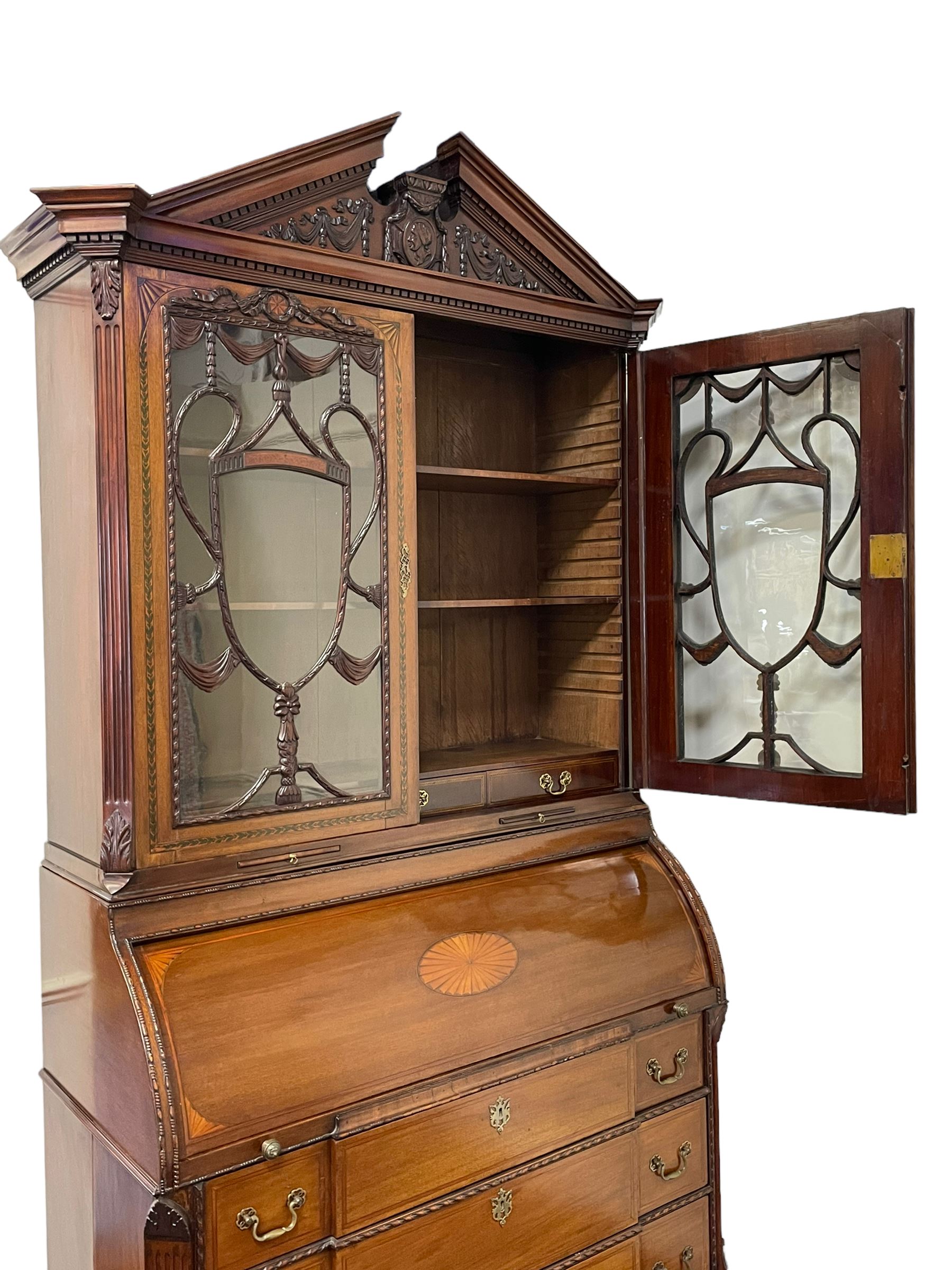 Large and impressive late 19th century Irish mahogany secretaire bookcase, moulded sloped arch dentil pediment decorated with central carved mask surrounded by linen festoons and trailing bellflowers, canted and fluted uprights with carved acanthus leaf upper and lower brackets, enclosed by two astragal glazed doors in the form of trophies with twist ribbon carved mouldings, inlaid with fanned spandrels and trailing leaf painted satinwood band, cylinder roll top inlaid with central fan motif and fanned spandrels, pull-out action revealing stepped fitted interior, central tambour roll cupboard flanked by two Composite pilasters and a combination of drawers and pigeon holes, fitted with three break-front drawers, matched canted and fluted corners with acanthus leaf carvings, on square tapering and fluted spade feet