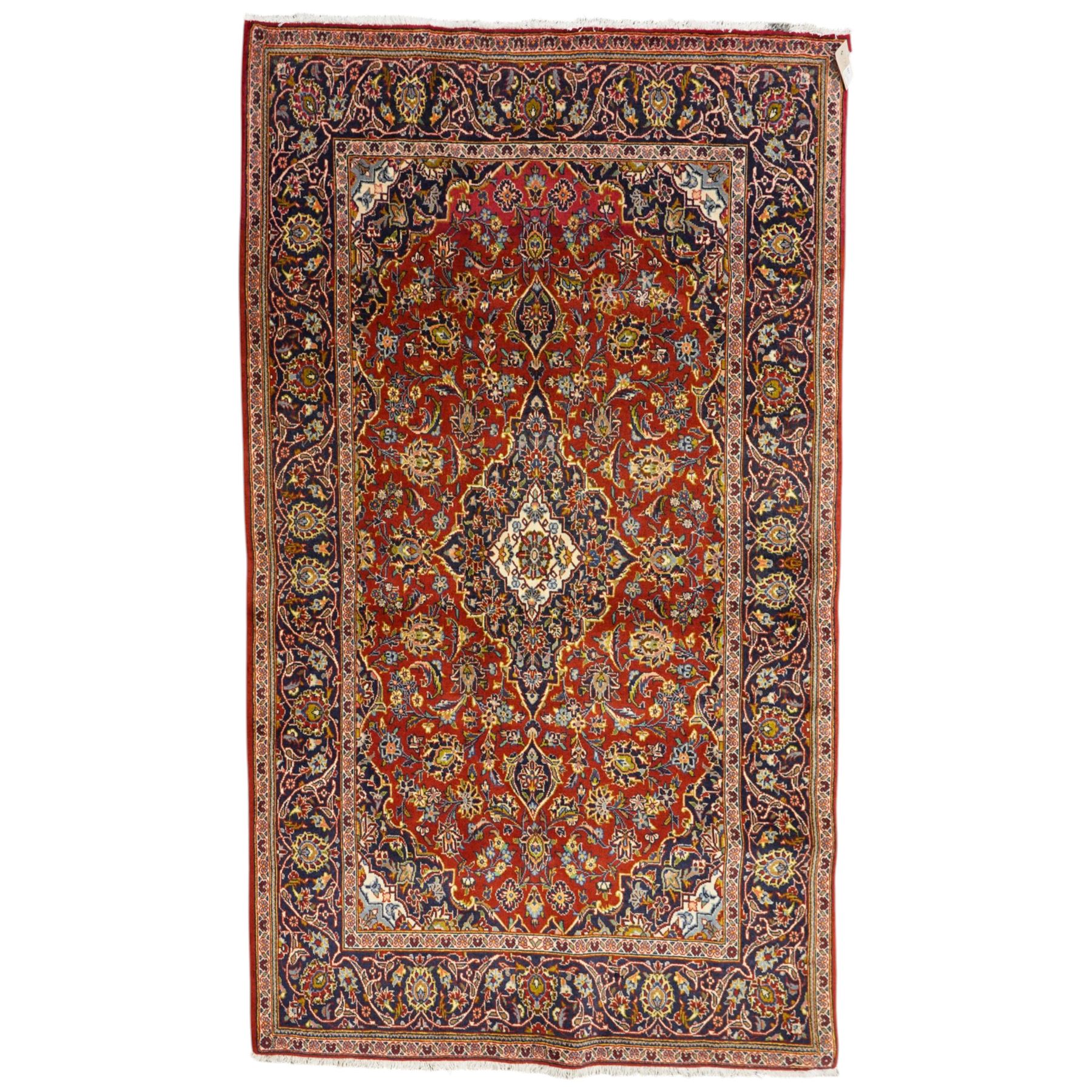 Central Persian Kashan hand-knotted rug, deep red field with central cusped ivory medallion with an all-over arrangement of scrolling floral vines, palmettes and flowering sprays within conforming spandrels, principal navy border with a continuous meandering vine of palmettes and stylized floral motifs, enclosed by multiple narrow guard borders, with cotton fringe ends 