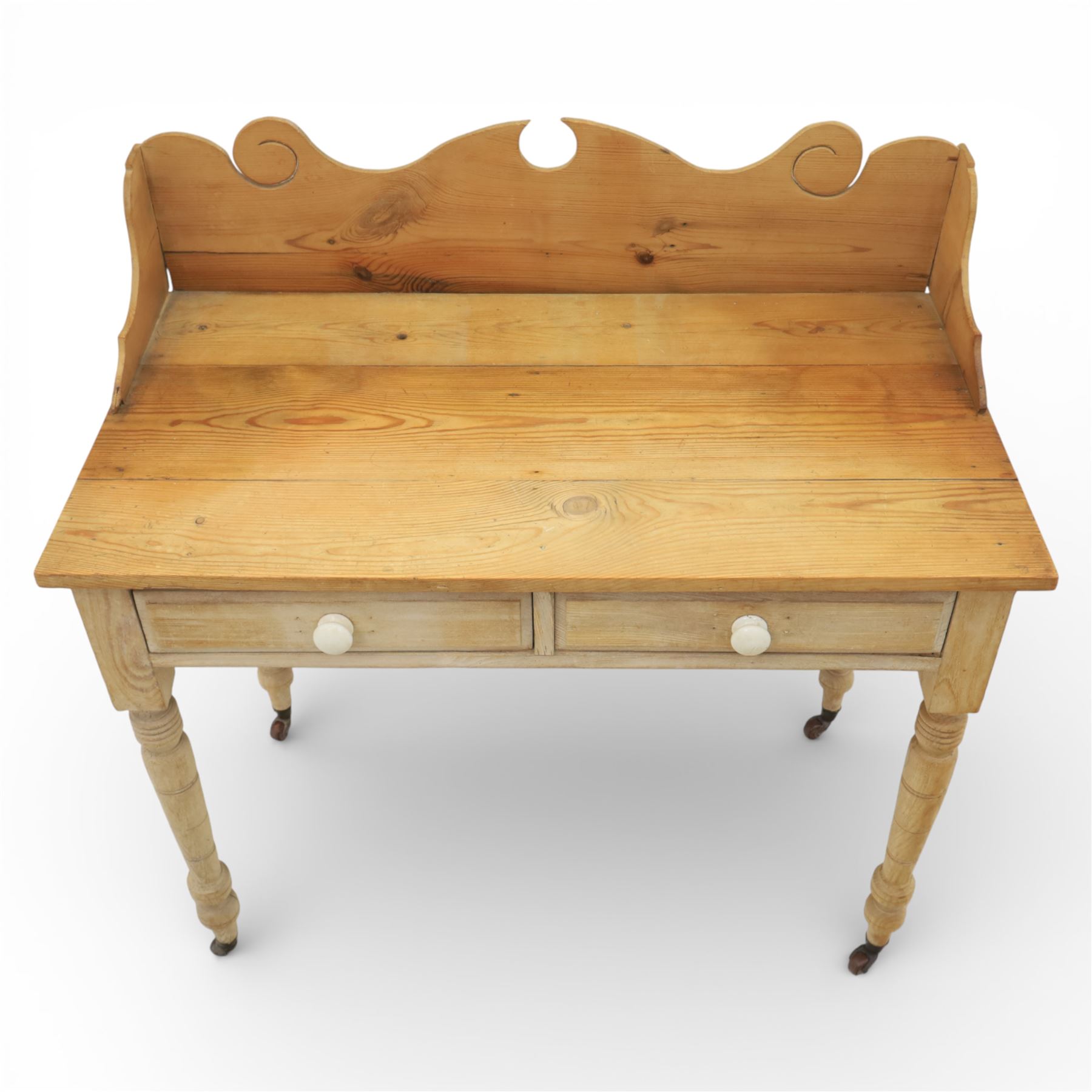 19th century stripped pine side table, the rectangular plank top with low shaped back rail incorporating scroll cut ends, fitted with two frieze drawers with ceramic knob handles, raised on turned supports terminating in castors