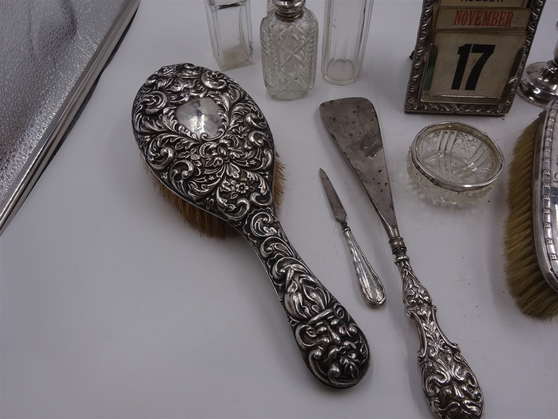 Group of silver, comprising Victorian silver mounted perpetual calendar with various date cards, hallmarked Henry Matthews, Birmingham 1900, silver mounted dressing table items, to include hair brush, with embossed decoration, pair of clothes brushes, and a shoe horn, together with a pair of silver mounted dwarf candlesticks, four cut glass jars/scent bottles with silver covers and a cut glass open salt with silver collar, all hallmarked 