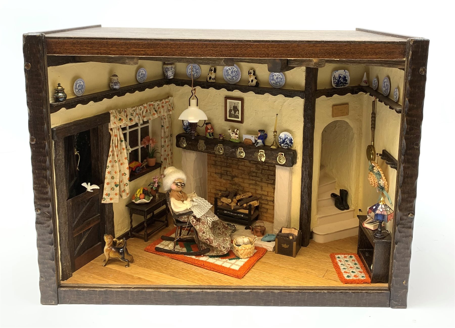 A 1/12th scale diorama of a cottage sitting room, depicting an elderly lady seated before a fireplace knitting, with dog and cat, in an interior furnished with blue and white plates, Staffordshire figures, toby jug, brass horse shoes, copper kettle, etc., H29cm L40cm D26cm.