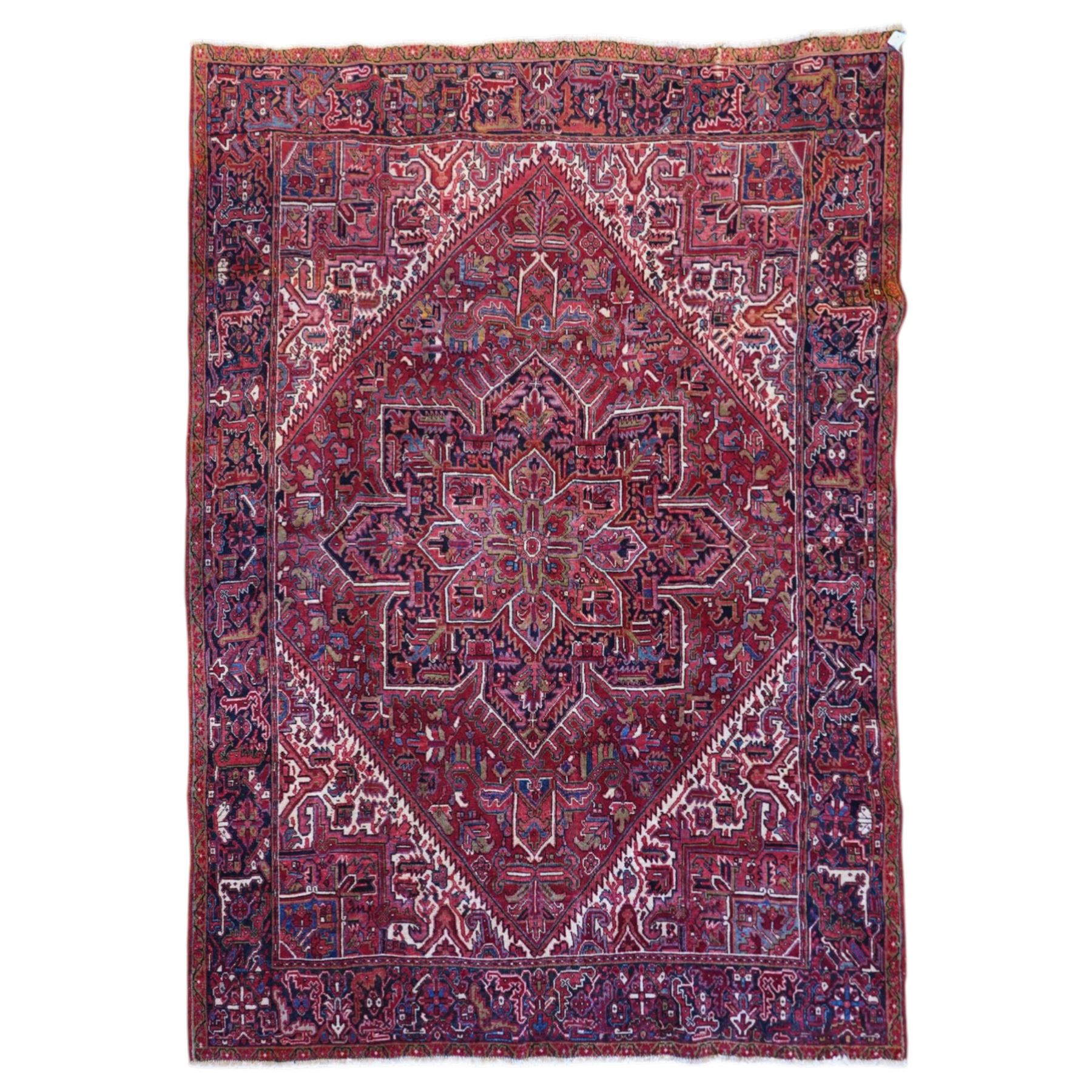North West Persian Heriz hand-knotted carpet, brick-red field with a large central angular star medallion with pendant motifs, surrounded by stylized geometric palmettes, serrated leaves and tribal devices, ivory corner spandrels similarly decorated, field enclosed by a dark indigo main border of stylized rosettes and angular vine motifs between multiple guard stripes, with cotton fringe ends  