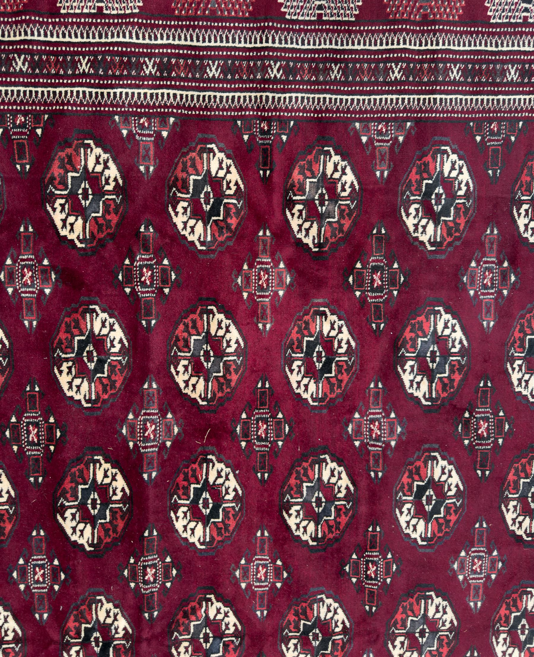 Northeastern Persian Yamut  hand-knotted carpet, the rich burgundy field with repeating rows of stepped gul motifs in cream and black, enclosed by multiple linear and geometric borders with hooked motifs and diamond medallions
