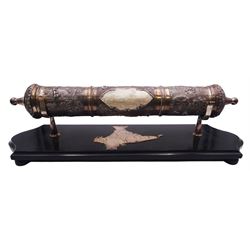 Late 19th/early 20th century Indian silver scroll holder, of cylindrical form with vacant cartouche and embossed with figures, animals and huts, upon a shaped ebonised stand with silver supports and silver panel of India