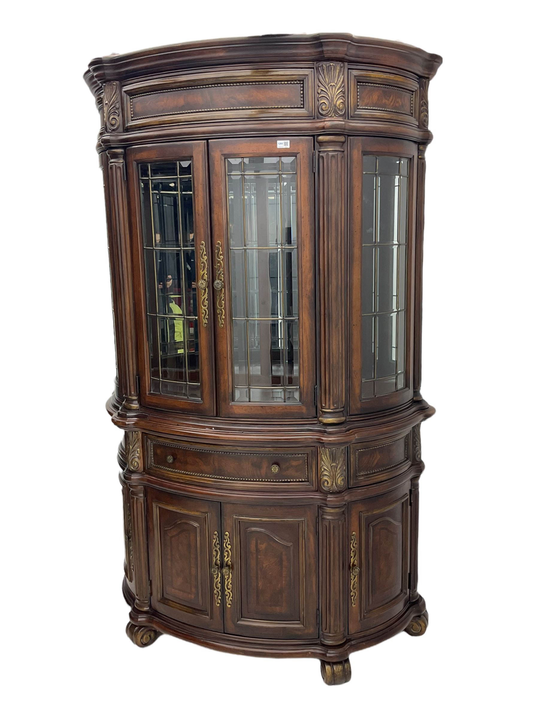  Kevin Charles American walnut demi lune display cabinet with illuminated interior, projecting cornice, two bevel edge glazed doors enclosing two adjustable shelves above single drawer and four cupboard doors with wine rack, carved scrolling supports