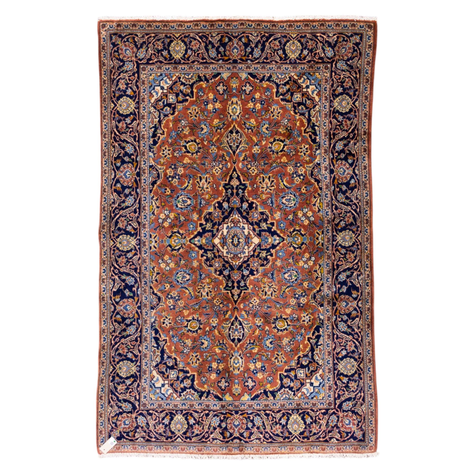 Central Persian Kashan hand-knotted rug,  terracotta field with central shaped ivory and dark blue medallion with palmettes, floral sprays and scrolling vine ornament, with conforming corner spandrels enclosed by a broad navy border of palmettes and flowering vine between multiple guard stripes, with cotton fringe ends 