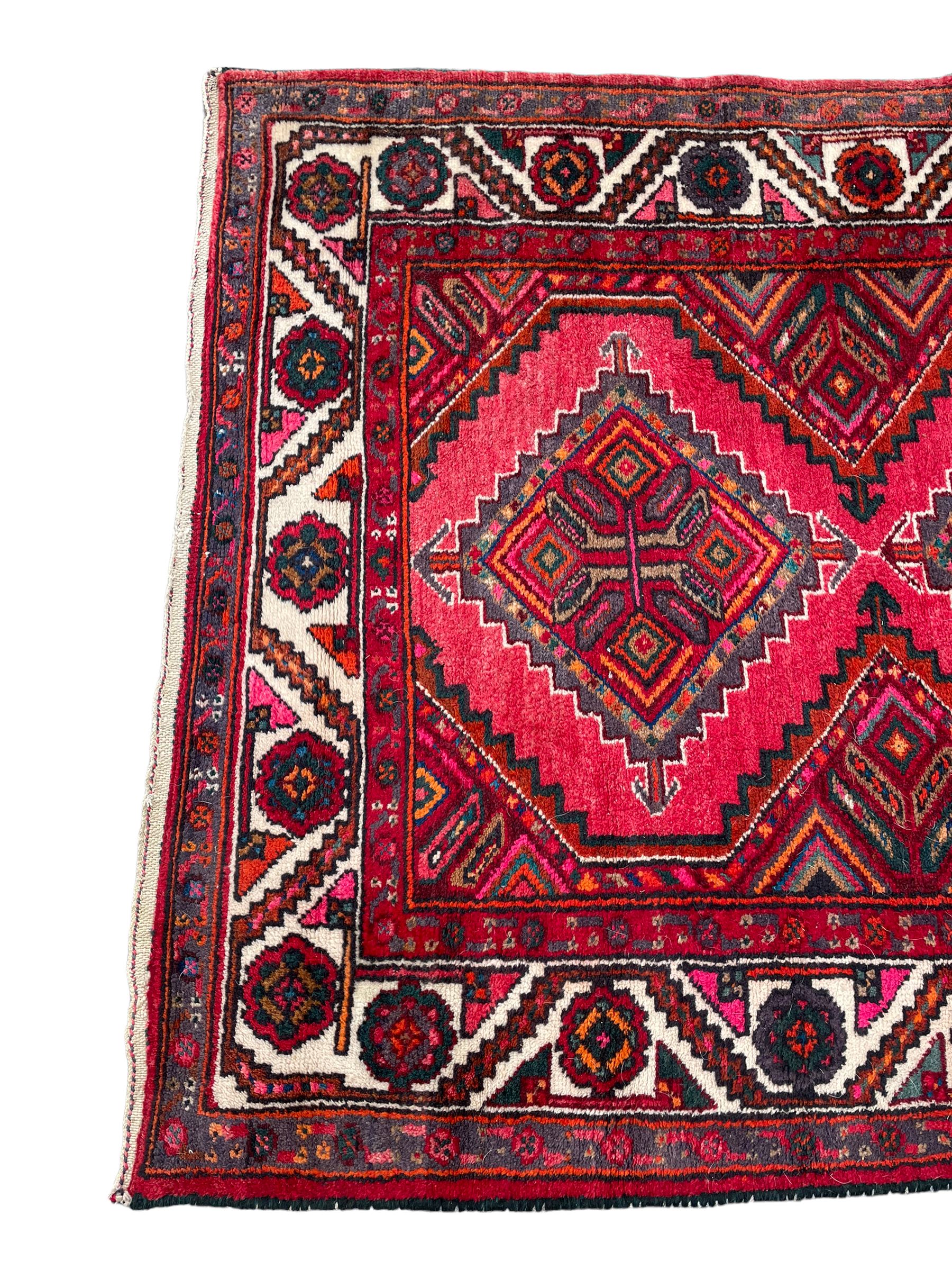 Persian Hamadan red ground rug, decorated with six stepped lozenge medallions, the guarded border with repeating stylised flower head decoration, with inscription to corner