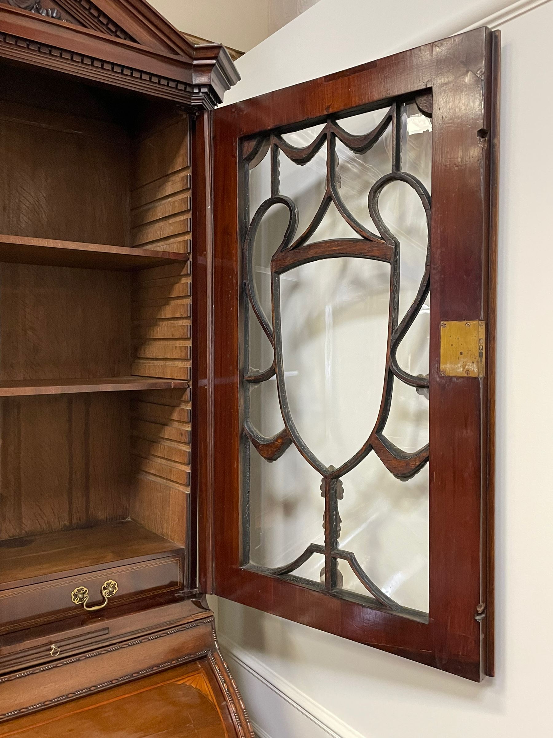Large and impressive late 19th century Irish mahogany secretaire bookcase, moulded sloped arch dentil pediment decorated with central carved mask surrounded by linen festoons and trailing bellflowers, canted and fluted uprights with carved acanthus leaf upper and lower brackets, enclosed by two astragal glazed doors in the form of trophies with twist ribbon carved mouldings, inlaid with fanned spandrels and trailing leaf painted satinwood band, cylinder roll top inlaid with central fan motif and fanned spandrels, pull-out action revealing stepped fitted interior, central tambour roll cupboard flanked by two Composite pilasters and a combination of drawers and pigeon holes, fitted with three break-front drawers, matched canted and fluted corners with acanthus leaf carvings, on square tapering and fluted spade feet