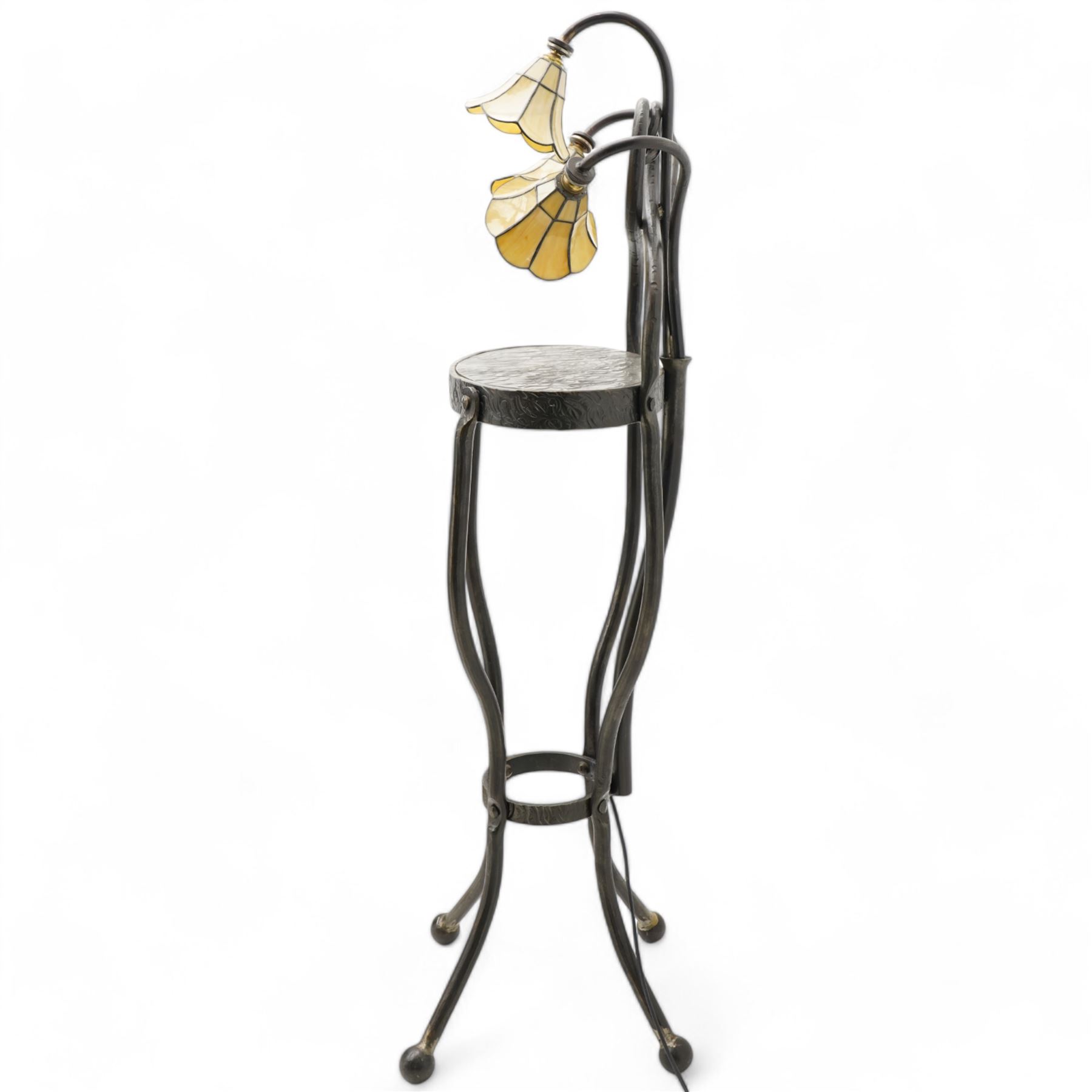 James Godbold - Art Nouveau blacksmith-made wrought iron floor standing lamp table, the round etched metal tabletop decorated with swirling foliage motifs, crowned by three Tiffany design amber glass shades mounted on curved iron stems, raised on four splayed supports united by circular stretcher and terminating in ball feet