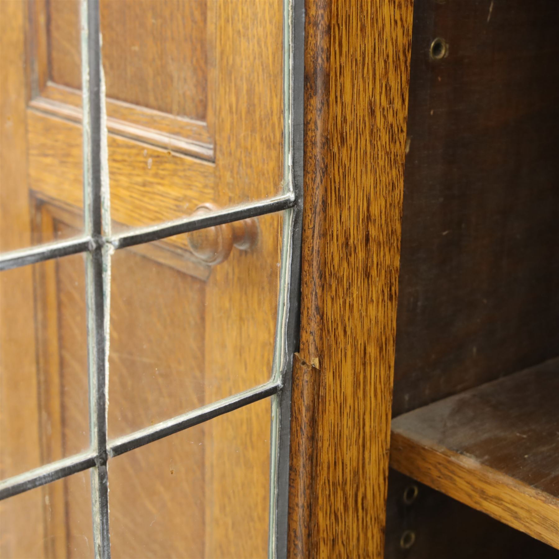 Edwardian oak glazed cabinet, moulded cornice above a panelled front and sides, fitted with lead glazed doors enclosing shelved interior, left side full length cupboard converted to enclose a gun cabinet, on a plain plinth base 