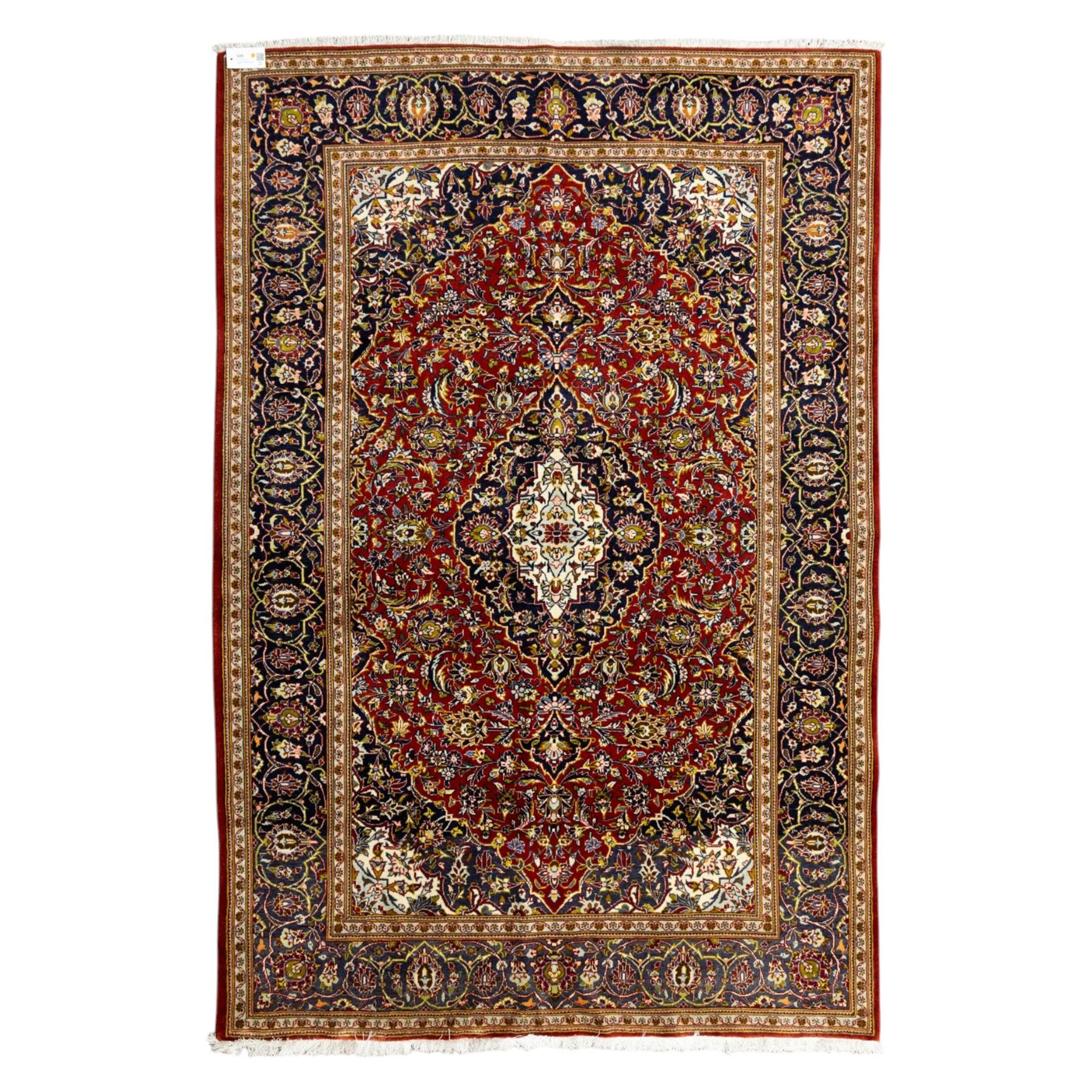 Central Persian Kashan hand-knotted carpet, crimson ground, with a central ivory shaped floral medallion with pendants, surrounded by dense scrolling foliate and floral motifs incorporating rosettes, palmettes and delicate vine work, enclosed by ivory spandrels and a dark navy border with repeating palmettes and meandering vines, flanked by floral guard stripes, with cotton fringes