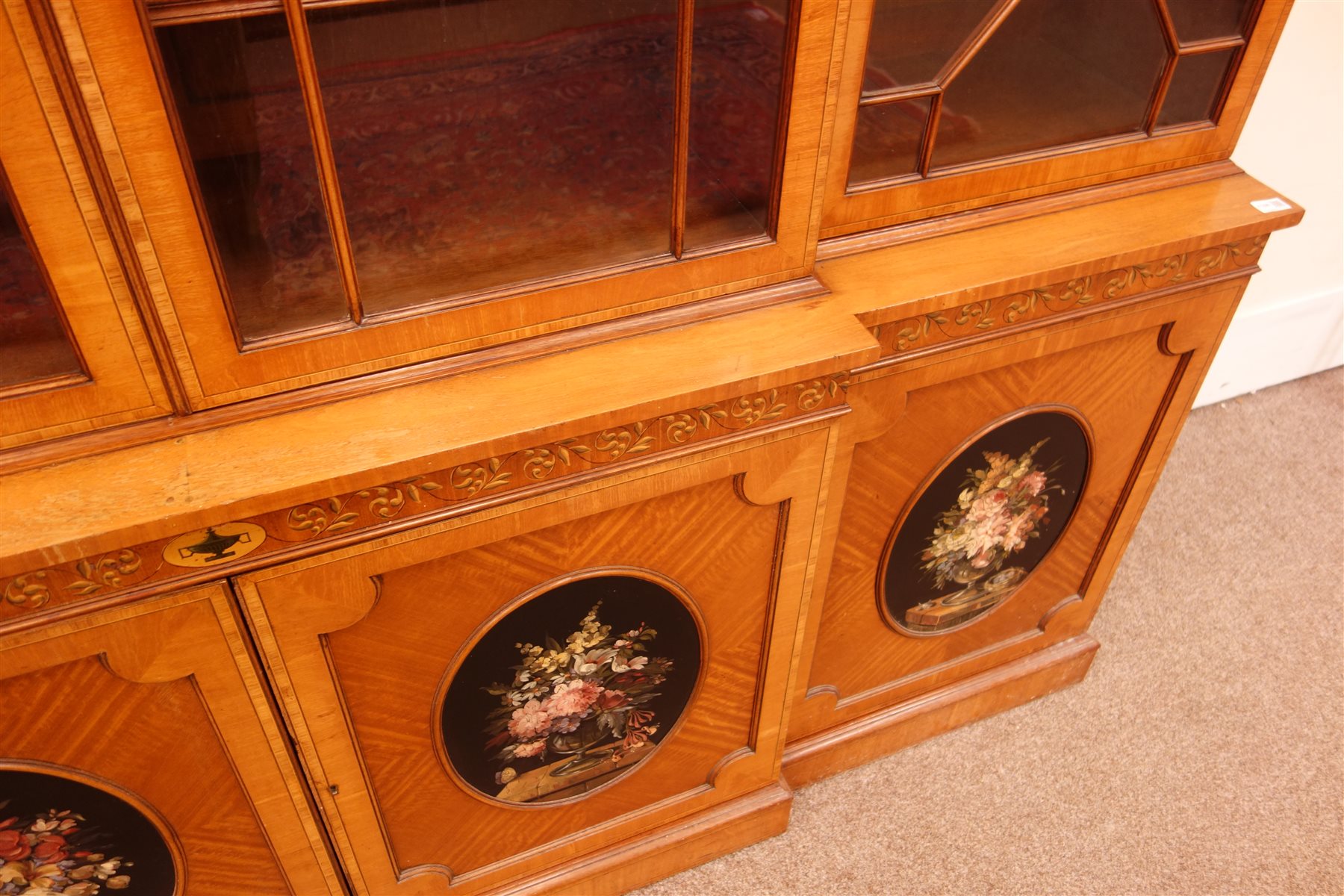 Sheraton Revival satinwood breakfront bookcase, frieze painted with scrolling leafage above four astragal doors with adjustable shelves, the base with four doors painted with floral sprays in oval panels, on a plinth base, W234cm, H235cm, D52cm