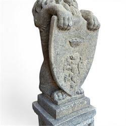 Pair of cast stone Heraldic lions, modelling in upright standing poses with flowing mane, each clasping an Armorial shield moulded with a crowned lion in their forepaws, raised on rectangular pedestals