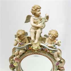  A late 19th century Sitzendorf porcelain mirror, the mirror plate of circular form set within a shaped surround encrusted with flowers and foliage, surmounted by three figures of cherubs and raised upon four scroll feet, with blue cross hatch mark beneath, H57cm.   