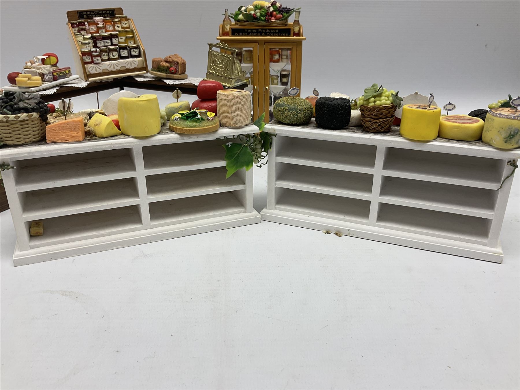 Collection of miniature dolls house grocery shop furniture and produce, comprising glazed corner counter with brass style cash register, shelved preserves unit, pies, cheese board and trays of meats, pair of white cheese counters, biscuit display with six tins, glazed preserves cabinet, pair of shop chairs and four further cheese boards complete with grapes and label flags