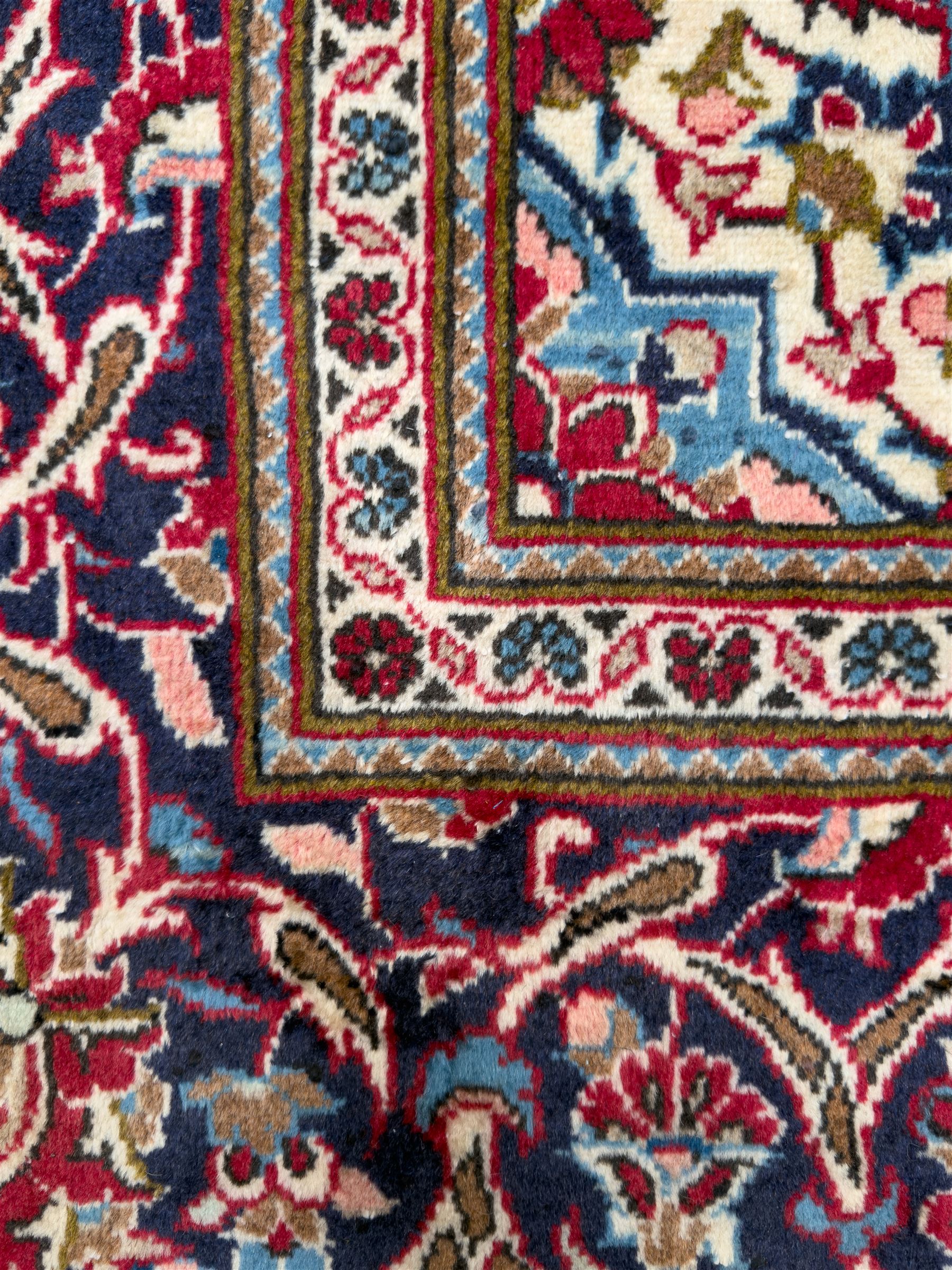 Central Persian Kashan hand-knotted carpet, the deep red ground with a central lozenge medallion in ivory and navy, surrounded by dense scrolling floral and vine motifs, navy spandrels and a wide border with stylised palmettes and flowerheads enclosed by multiple guard stripes