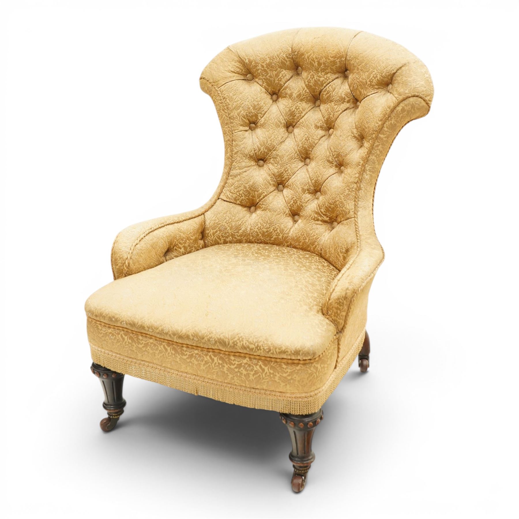 Victorian Aesthetic Movement walnut framed nursing chair, the fan back and sprung seat upholstered in buttoned foliate patterned gold damask fabric with fringing, raised on beaded tapering fluted supports with ceramic castors