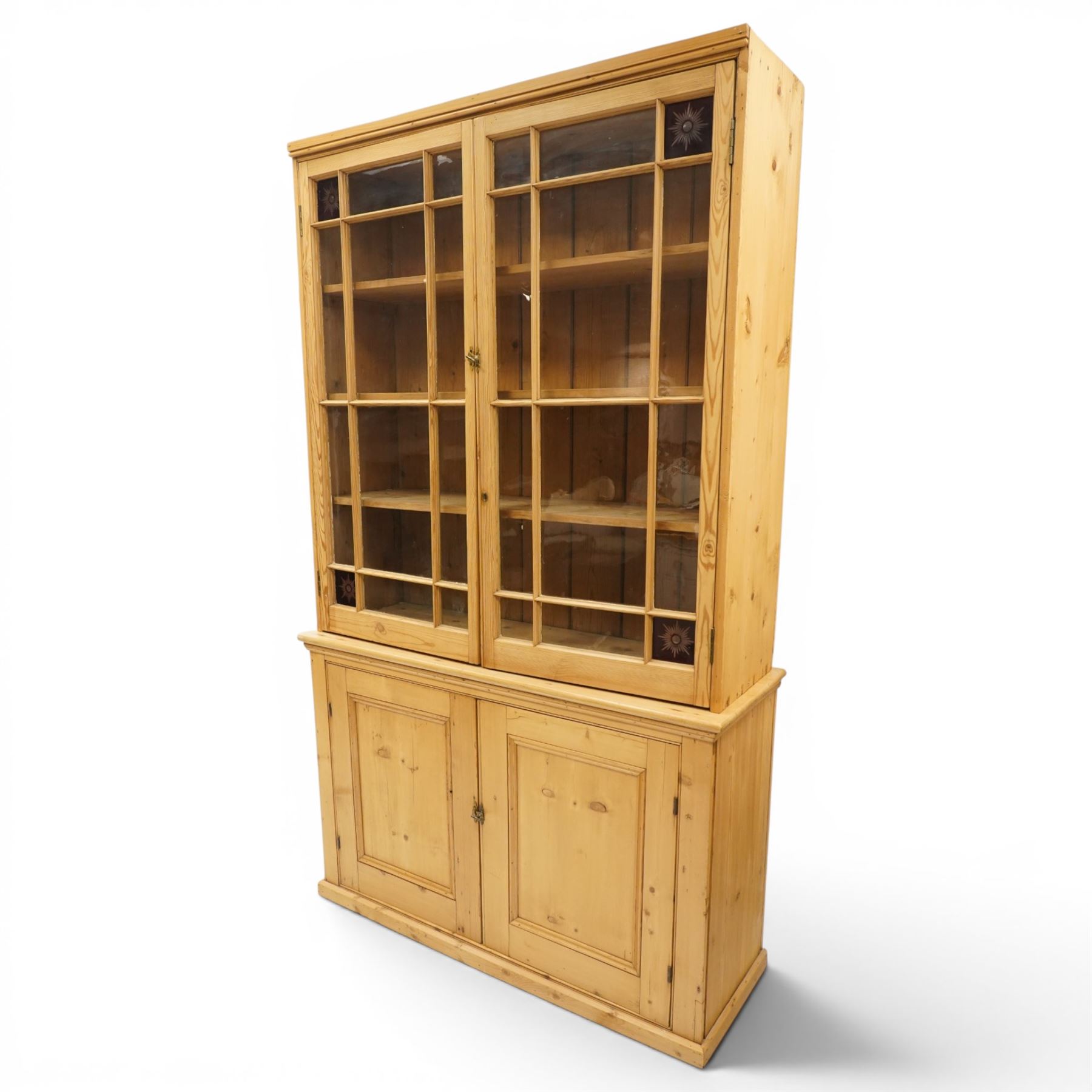 19th century pine glazed bookcase cupboard, twin astragal glazed doors with stained glass corner panels, enclosing fixed shelves, the lower section fitted with two panelled cupboard doors enclosing single shelf, on plinth base