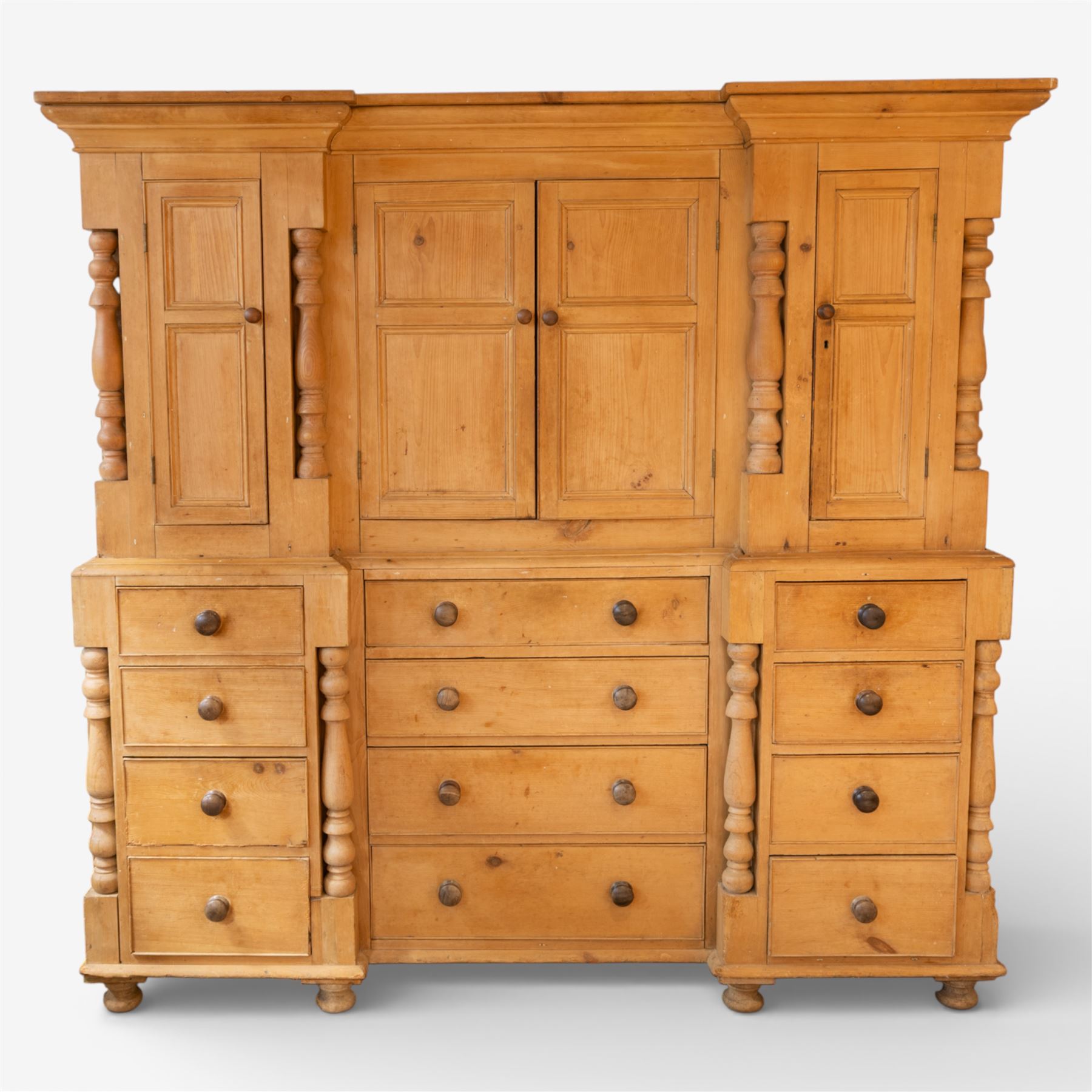 Large Victorian stripped pine reverse breakfront housekeeper’s cupboard, the moulded cornice above a central pair of panelled cupboard doors flanked by single cupboard doors and divided by turned baluster pilasters, the lower section fitted with an arrangement of twelve drawers with knob handles, raised upon bun feet