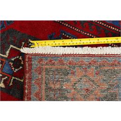 North East Persian Balochi hand-knotted runner, crimson field decorated with three vertical columns of repeating octagonal guls, each enclosing angular geometric motifs in deep indigo, ivory, and soft brown tones, guls linked by stepped diamond and cruciform devices, main border of stylized geometric floral motifs and angular vines, flanked by multiple guard borders, with cotton fringe ends 