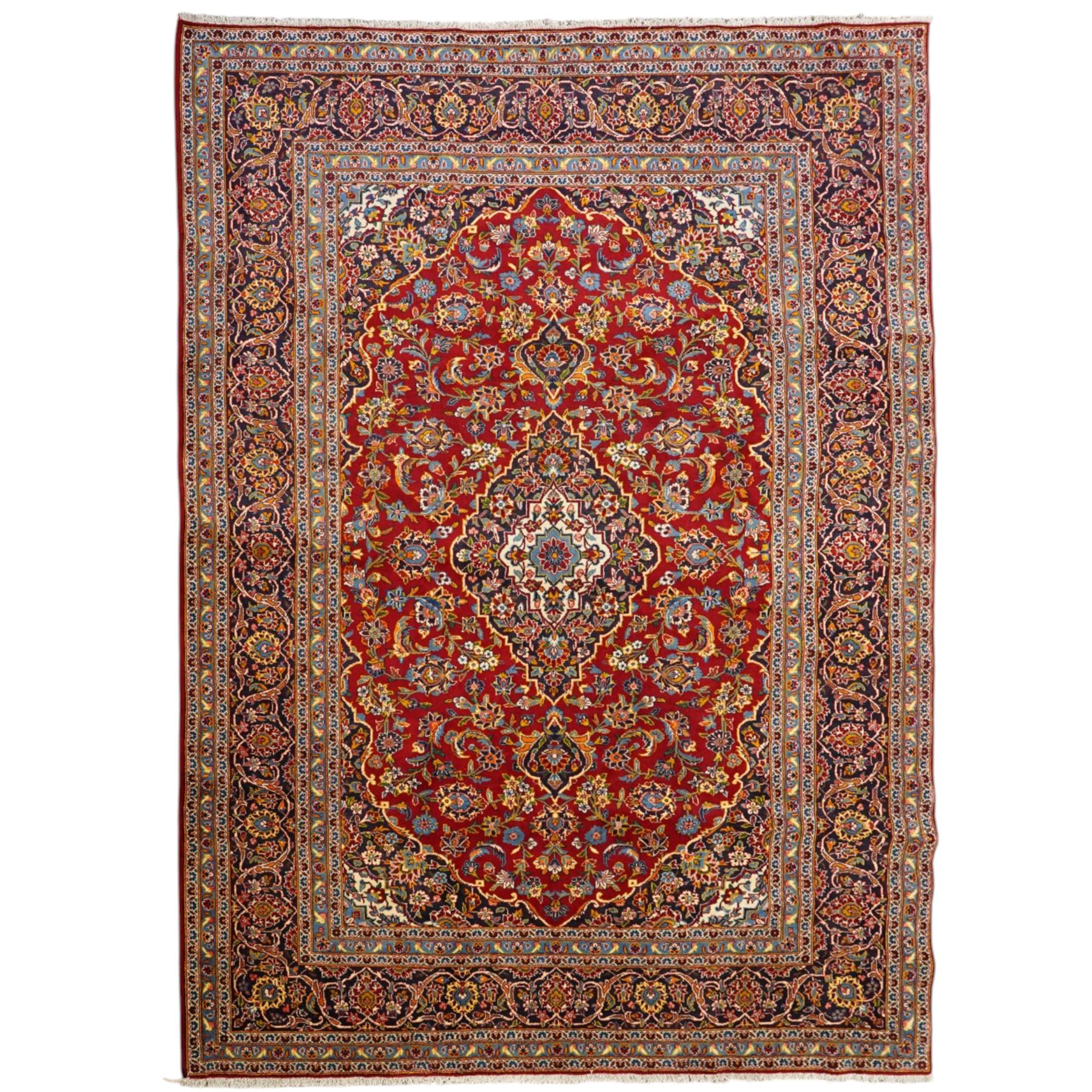Central Persian Kashan hand-knotted carpet, crimson field with central cusped medallion in ivory, pale blue and indigo, issuing a network of scrolling arabesques, palmettes, and floral sprays, with conforming spandrels, main border of scrolling vinery and stylized palmettes, framed by multiple guard borders, with cotton fringe ends 