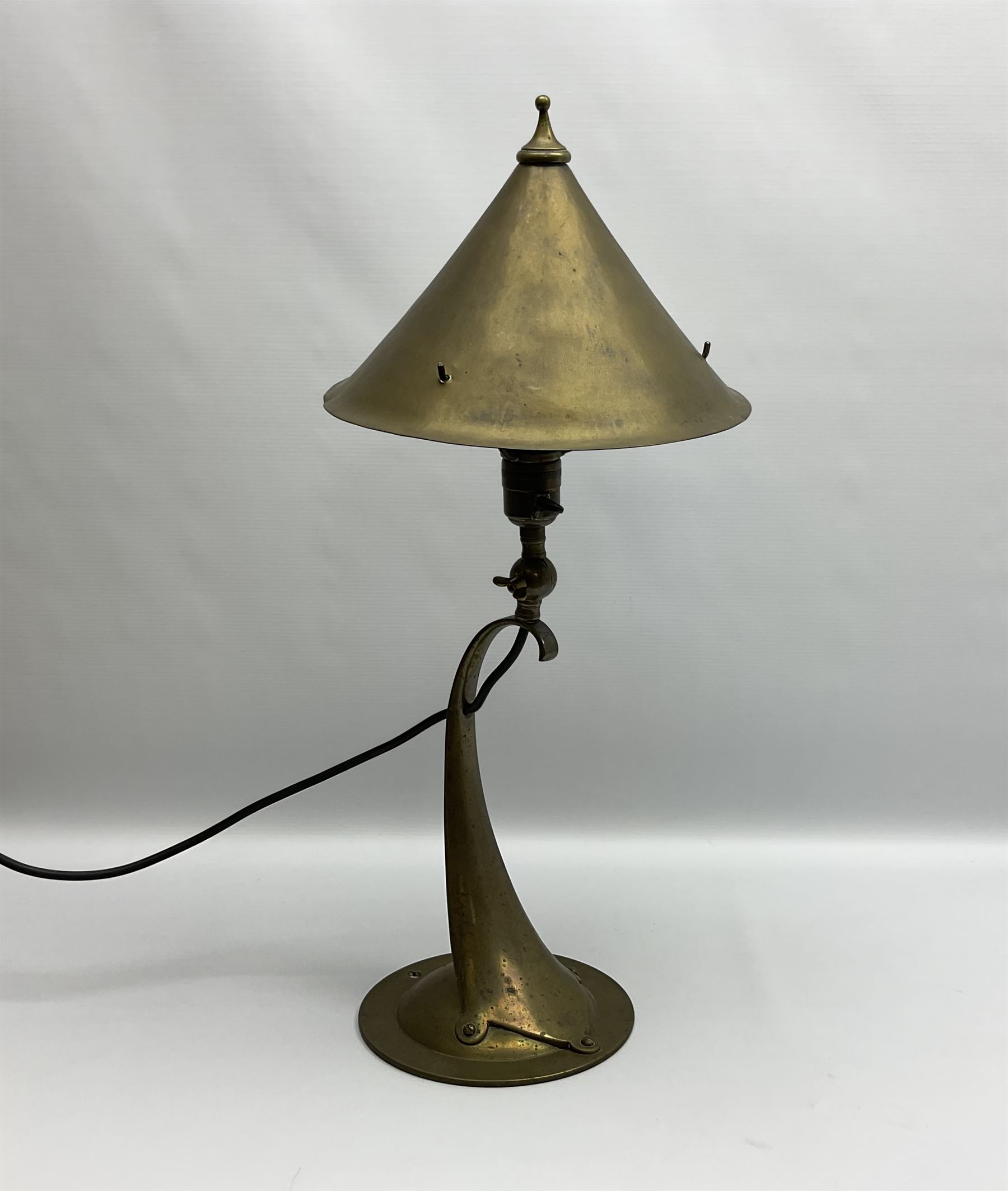 Arts & Crafts Jesson, Birkett & Co, brass table lamp or wall light, designed by Thomas Birkett, the tapering stem with adjustable ball pivot supporting a conical shade with tapering final with ball terminal, upon a circular domed base pierced for wall mounting, shade positioned upright overall H57cm

Often misattributed to Arthur Stansfield Dixon for Birmingham Guild of Handicraft, this particular lamp was designed by Thomas Birkett.
In the late 1890's Birkett worked as a metalwork at the Birmingham Guild, before later joining the Faulkner Bronze Company which went on to become Jesson, Birkett & Co in 1904. The lamp features in lighting catalogues from the firm under the model no E490.

