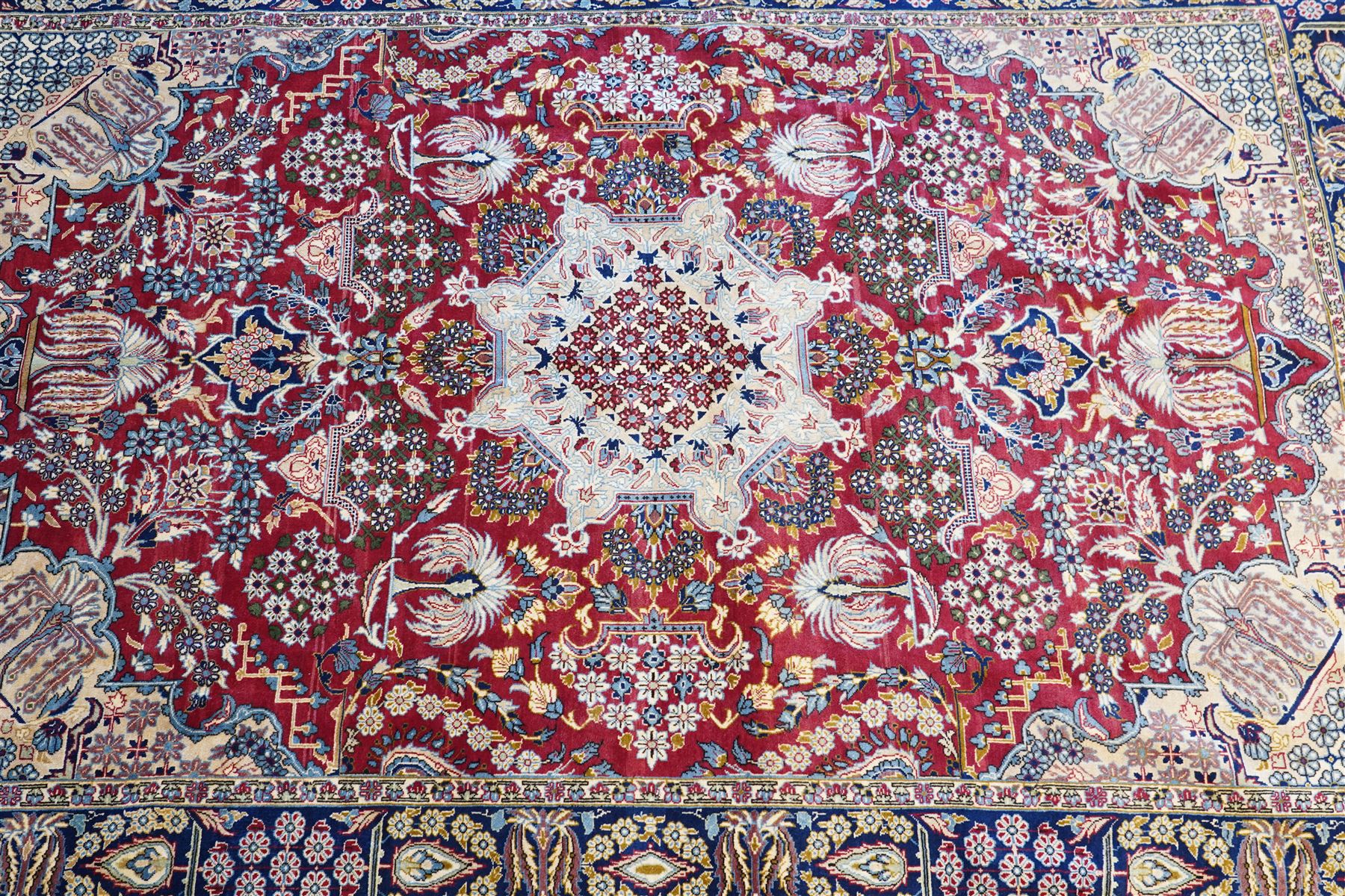 Central Persian Najafabad hand-knotted carpet, crimson field with a central elaborate cusped medallion with radiating floral motifs, surrounded by finely drawn scrolling vines incorporating stylized palmettes and rosettes, flowering plants and vase motifs, ivory spandrels similarly decorated enclosed by a dark indigo main border of floral arabesques between multiple guard stripes, with cotton fringe ends 