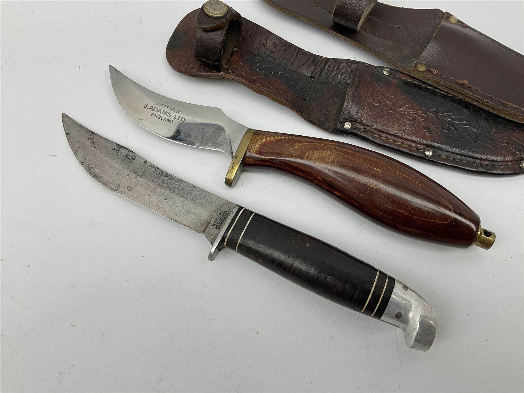 Western Boulder Colorado hunting knife the 11.5cm curving blade with maker's name to the ricassso, leather strip bound grip and aluminium pommel, in oak leaf decorated leather sheath L24cm; and J. Adams Sheffield hunting knife with curving blade and laminated wooden grip, in leather sheath (2)