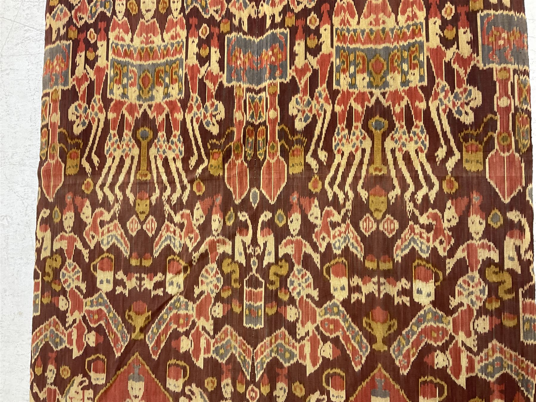 Ikat Man’s Cloth (Hinggi Kombu), East Sumba, Indonesia, woven in red, blue and yellow-brown warp ikat, constructed of two mirroring long panels, the motifs arranged in four bands depicting warrior figures astride rearing horses, high-ranking figures and guardians within geometric borders, on carved wooden wall hanger, L295cm
