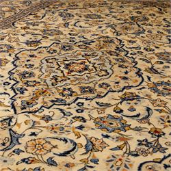 Central Persian Kashan hand-knotted carpet, ivory ground with central stepped floral medallion surrounded by scrolling vine work, palmettes, and stylized blossoms, field enclosed by a multi-border arrangement, wide main border decorated with repeating floral cartouches and foliate motifs, flanked by narrow guard stripes, with cotton fringe ends