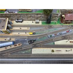 Table top 'N' gauge continental scenic layout as a split level town with various loops of track, station with numerous platforms, engine shed, assorted buildings, roads with motor vehicles and figures, tunnels, trees, river with bridge, tram depot etc 122 x 52cm