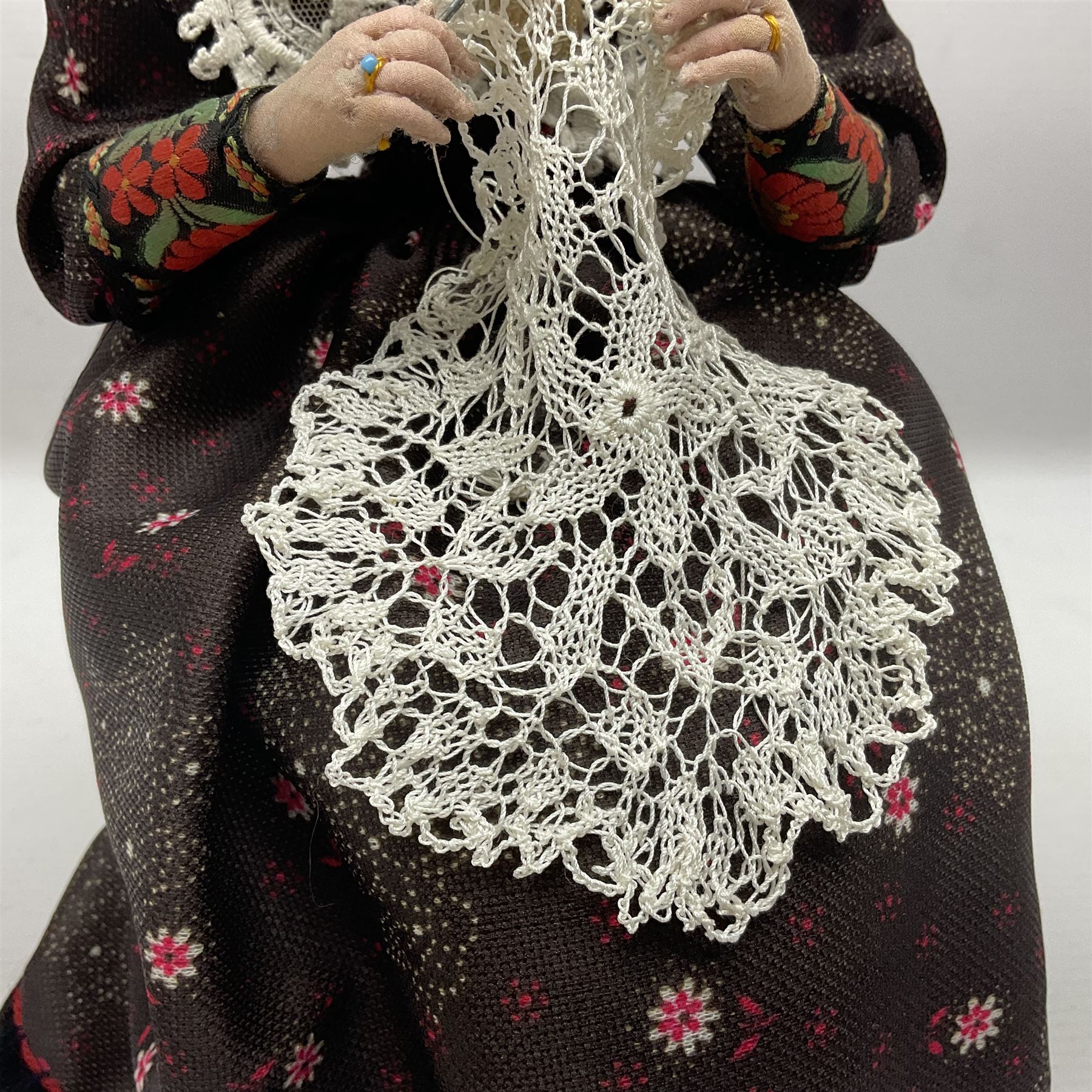 Anna Meszaros Hungary - hand made needlework figurine 'Old Lady' seated on a stool wearing a lace trimmed black/white/red floral full length dress and hat, working on a piece of lace H30cm  Auctioneer's Note: Anna Meszaros came to England from her native Hungary in 1959 to marry an English businessman she met while demonstrating her art at the 1958 Brussels Exhibition. Shortly before she left for England she was awarded the title of Folk Artist Master by the Hungarian Government. Anna was a gifted painter of mainly portraits and sculptress before starting to make her figurines which are completely hand made and unique, each with a character and expression of its own. The hands, feet and face are sculptured by layering the material and pulling the features into place with needle and thread. She died in Hull in 1998.