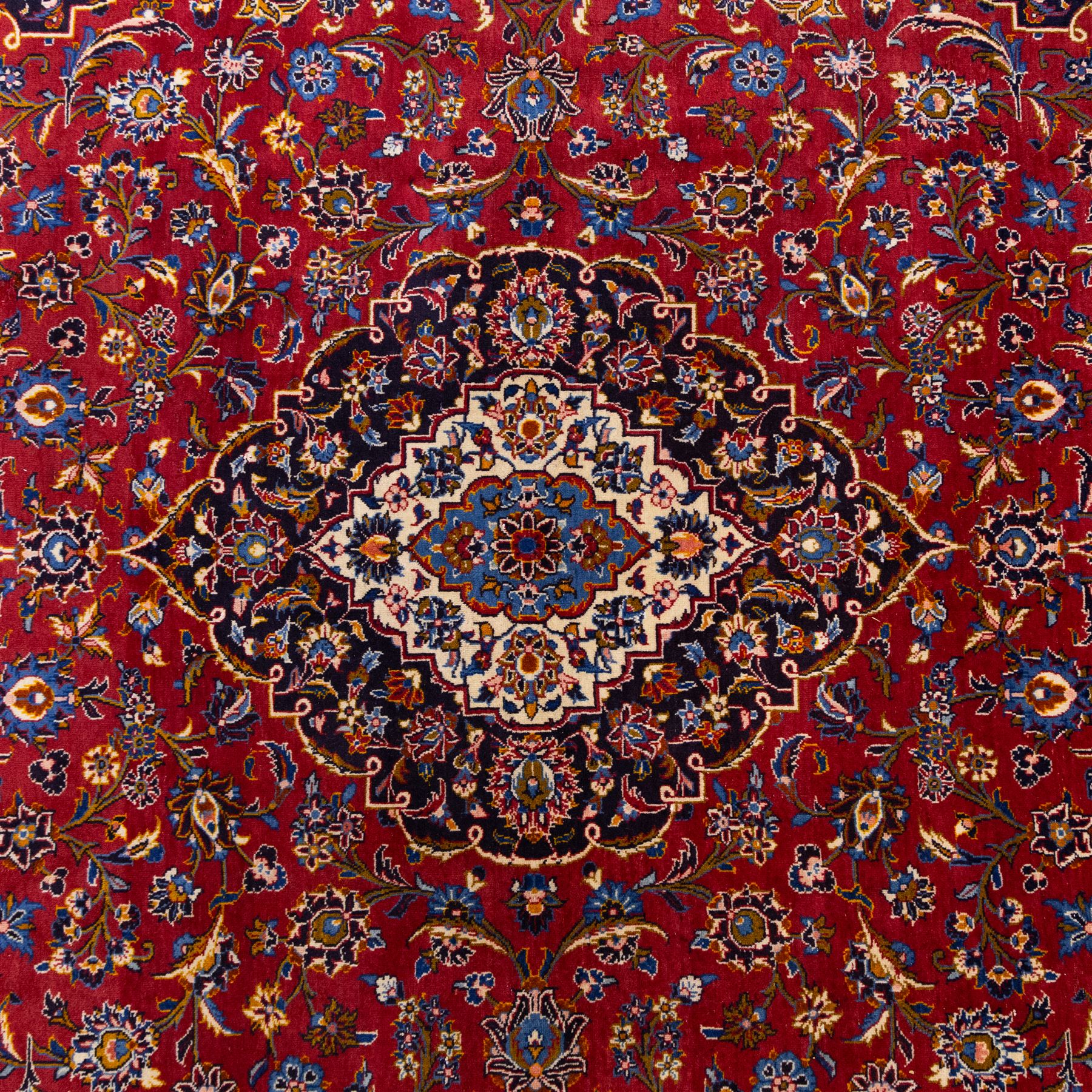 Central Persian Kashan hand-knotted carpet, rich crimson red field with a central cusped medallion with finely drawn floral sprays surrounded by dense scrolling vine work and palmettes, framed by a broad indigo ground border of flowering arabesques flanked by multiple guard stripes, in a palette of red, blue, ivory, sage green, and soft ochre tones, with cotton fringe ends    