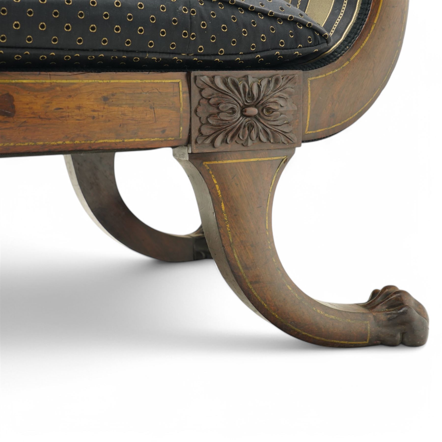 Regency brass inlaid rosewood chaise lounge or settee, shaped back over rolled head and foot, upholstered in black and gold striped fabric with rectangular squab cushion and bolster cushion, the rolled terminals carved with acanthus leaf roundels, lower rectangular foliate carved panels, on out splayed supports with paw carved feet 