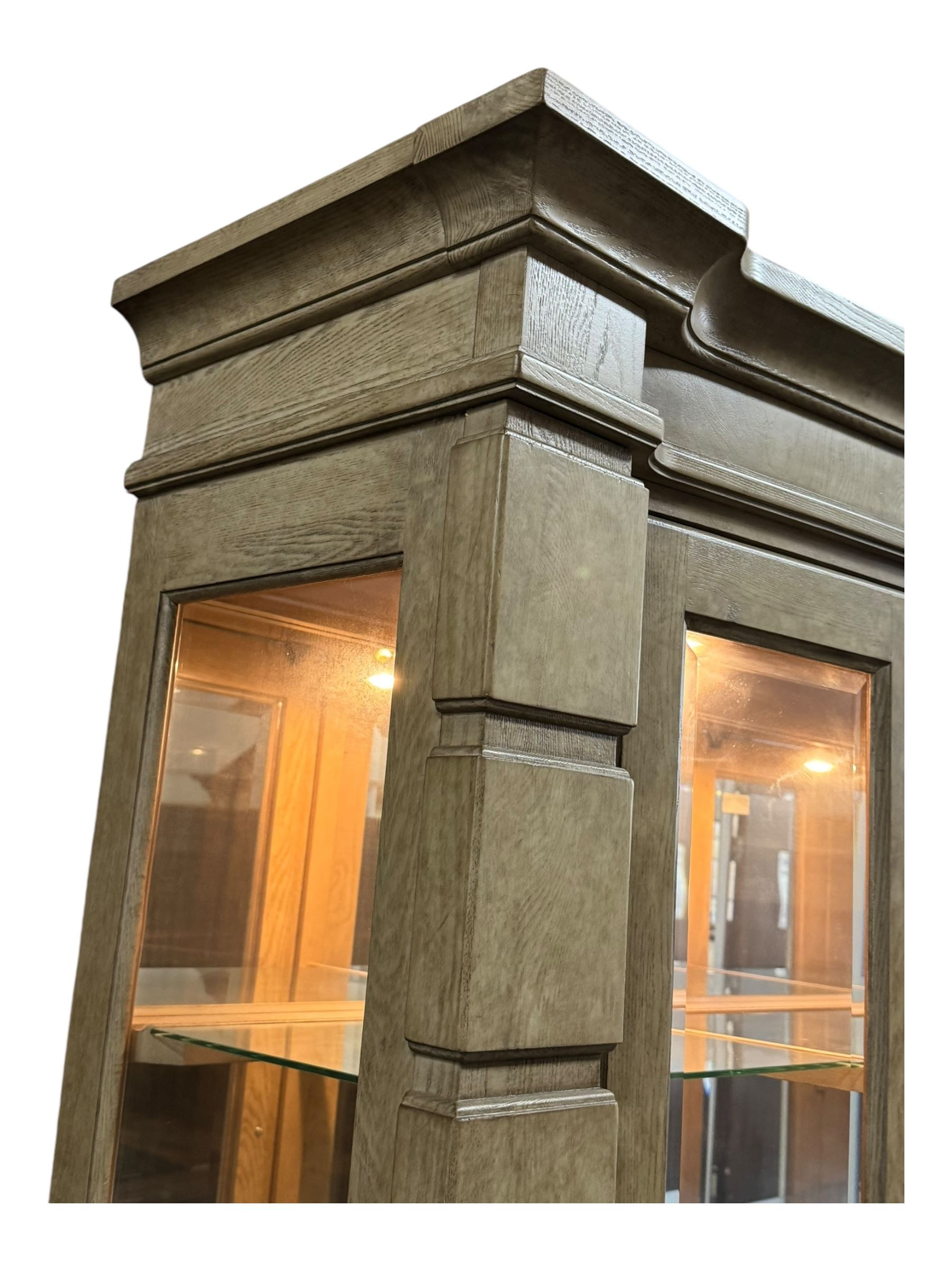 Brunswick - washed oak finish illuminated display cabinet, upper section with four glass-panelled doors enclosing glass shelving, base fitted with two drawers over four paneled cupboard doors, each fitted with distinctive round metal handles, raised on plinth base