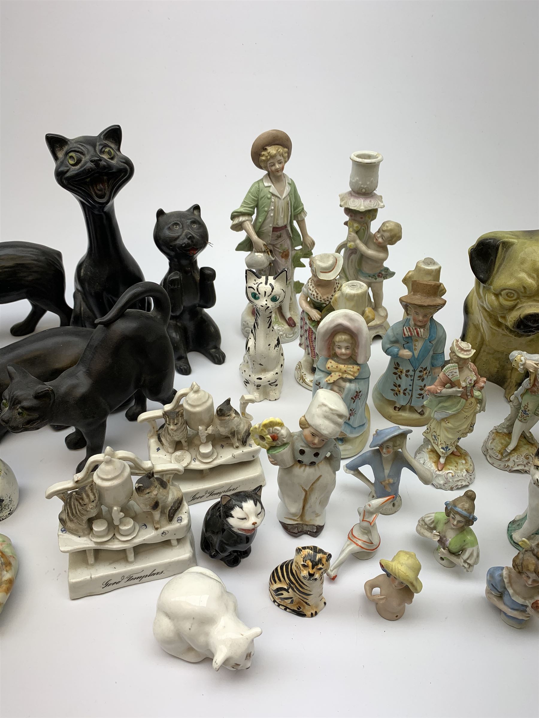 A collection of various ceramic figurines, to include pair of small 19th century Staffordshire cats, Royal Doulton cat, Coalport cat, pair of 19th century pug dogs, two fairings inscribed Good Templars, two Bonzo dog peppers, a number of pin cushion dolls, various 20th century and later black pottery cats, Victorian bisque figures, etc. 