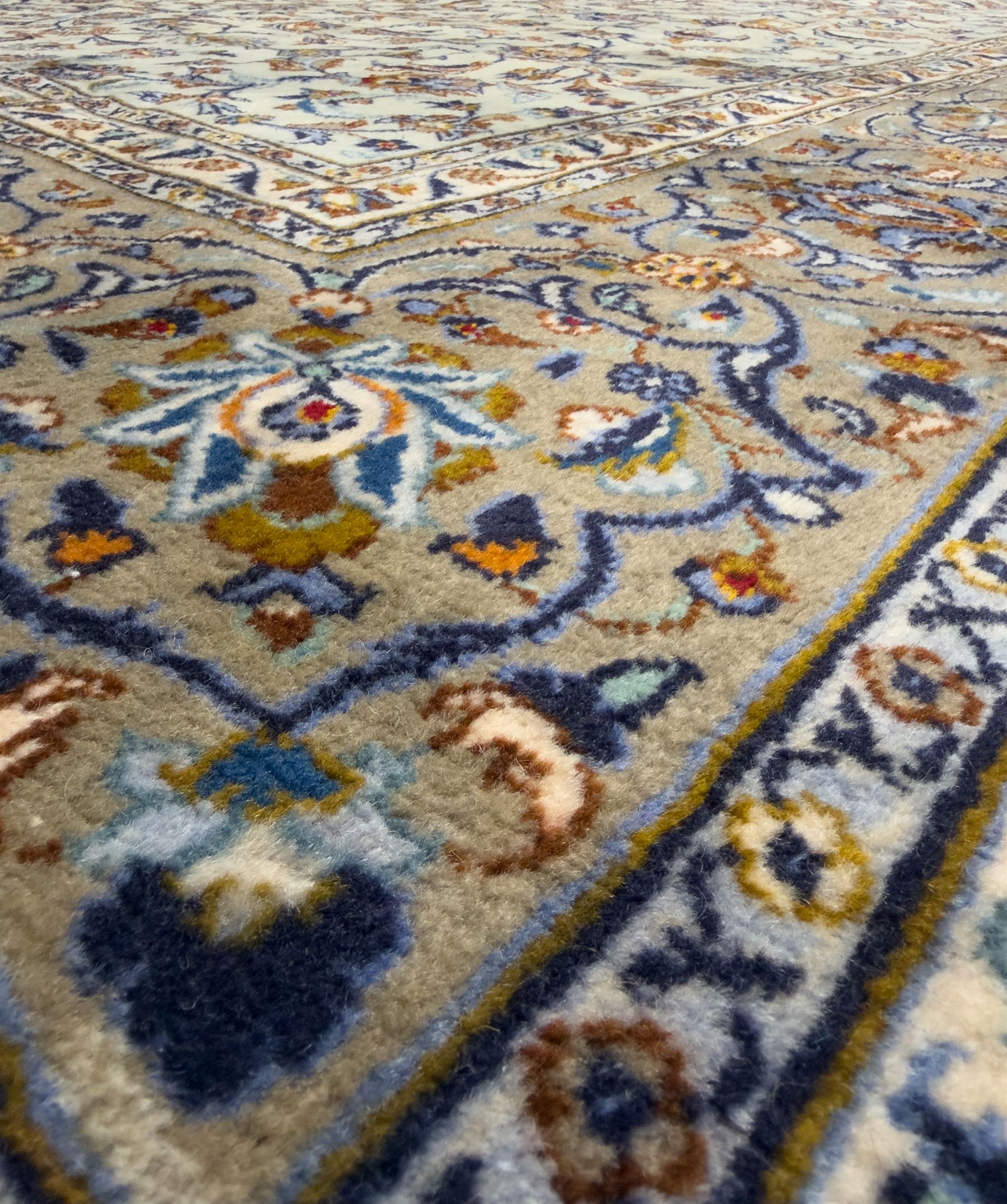 Central Persian Kashan hand-knotted carpet, the ivory ground intricately decorated with scrolling vines, palmettes, and stylised flowerheads in soft blue, gold and brown tones, enclosed by a broad indigo border with repeating floral motifs and multiple guard stripes