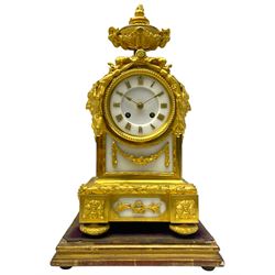 French late 19th century 8-day gilt ormolu mantle clock on a padded base – with urn surmount and floral garland crest on lobed disc cast feet, white marble two-part dial and panel beneath, dial with applied Roman numerals and gilt spade hands, Parisian rack striking movement, striking the hours and half hours on a bell. With pendulum and key.  