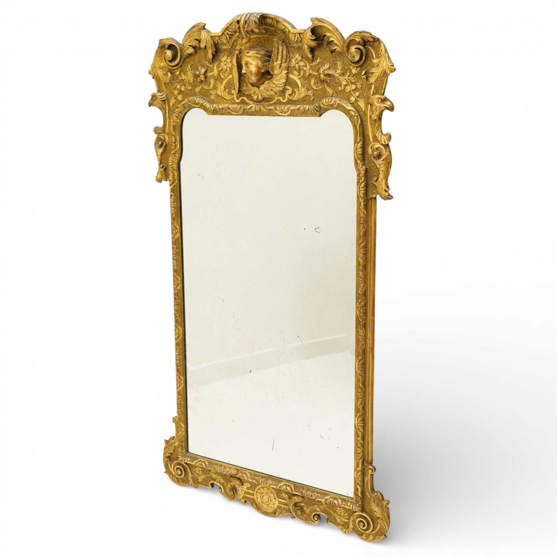 In the manner of Thomas Johnson - circa 1900 Georgian Revival giltwood wall mirror, the shaped rectangular plate within a foliate and scallop carved slip and richly ornamented frame, surmounted by a pierced cartouche crest centred with a protruding relief carved winged cherub mask and issuing scrolling acanthus and floral ornament, the sides with eagle masks over acanthus scrolls, terminating in a shaped apron with moulded rosette and spiralling acanthus leaves