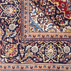 Central Persian Kashan hand-knotted rug, rich red field with a central lobbed medallion surrounded by finely drawn scrolling floral sprays, palmettes, and rosettes, enclosed by a broad dark blue floral border with multiple guard stripes and cotton fringe ends 