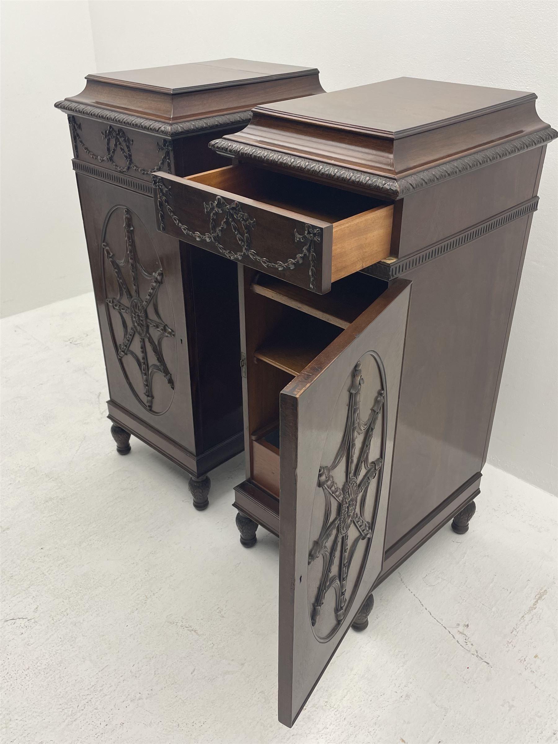 Pair early 20th century Hepplewhite style mahogany pedestal cabinets, cavetto moulded top with foliate carved edge over secret frieze drawer with applied trailing floral and ribbon decoration, each with button release underneath, oval panelled door with trailing harebell and central flower head motif decoration enclosing shelves, fitted cutlery drawer and lined cellarette drawer, on turned acanthus carved feet