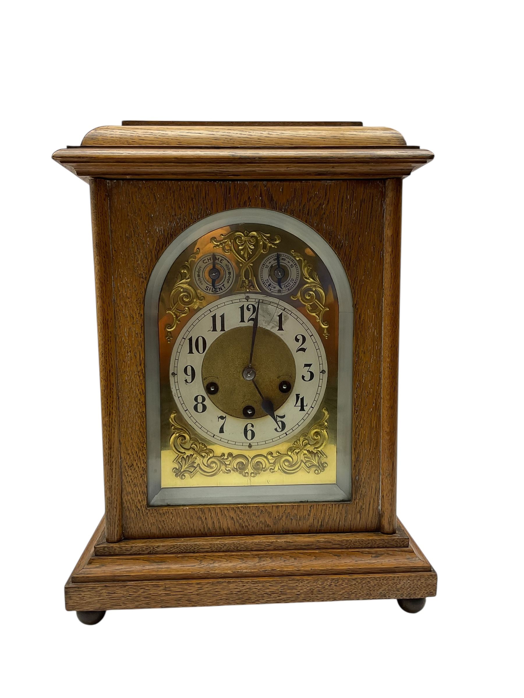 Jungans-German Edwardian 8-day oak cased mantle clock, with a stepped flat top and glazed door on a shallow plinth with bun feet, brass dial with cast spandrels, silvered chapter with Arabic numerals, chime/silent and pendulum regulation dials, three train spring driven movement sounding the quarters on gong rods and the hours on a coiled gong. With pendulum.