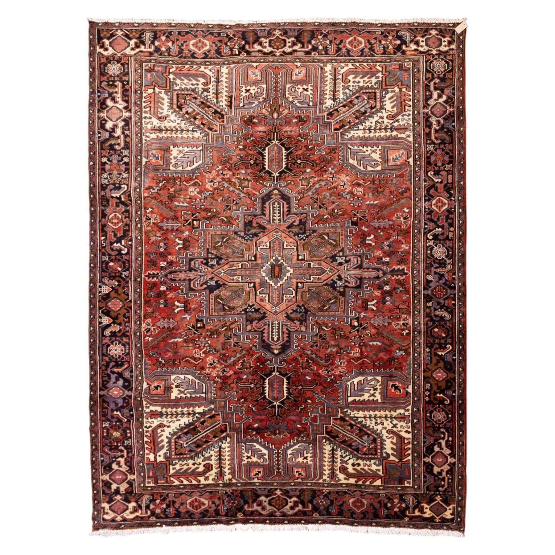 North West Persian Heriz hand-knotted carpet, madder red field with central stepped geometric medallion with radiating angular motifs and serrated outlines, and stylized floral and tribal devices framed by a broad border of repeating angular palmettes and rosettes flanked by multiple guard stripes, with cotton fringe ends 