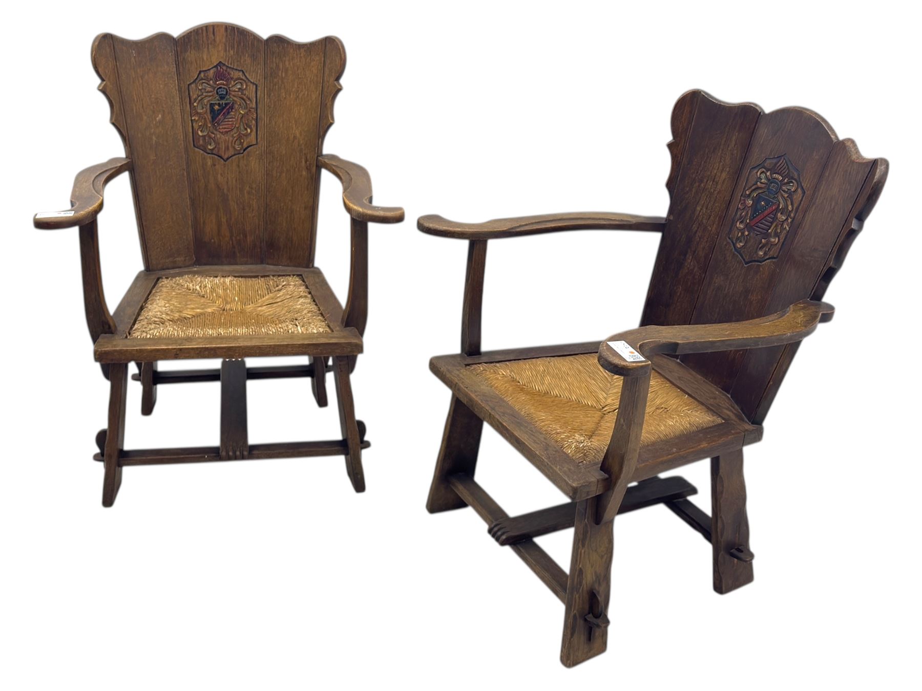 Pair of Arts and Crafts oak armchairs each with shaped backrest centred by polychrome painted crest, rush seat over straight square legs united by shaped central stretcher