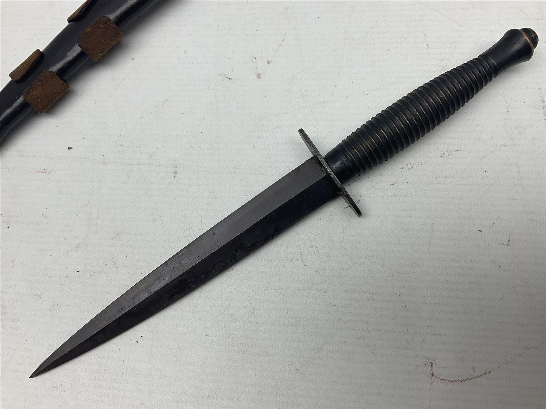 Third Pattern Commando Knife or Dagger with 17.5cm flattened diamond section blade and regulation black painted ribbed copper hilt with brass pommel nut; stamped William Rodgers Sheffield England to the cross-piece; in black painted brass mounted leather scabbard L34.5cm overall