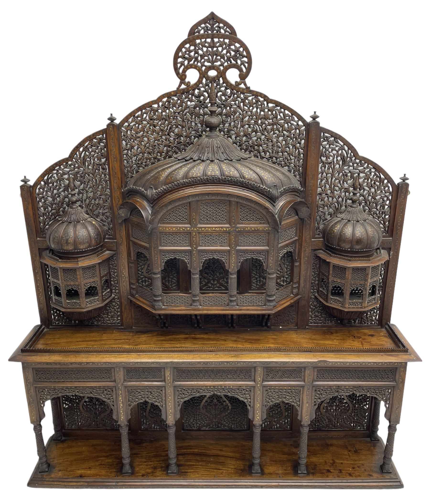 Late 19th century Indian Burmese carved and pierced hardwood and brass inlaid wall shelf, the shaped back profusely decorated with trailing and interlaced foliage, with projecting architectural structures, the central balcony with canopy top and five open windows with arched apertures on columns, two flanking five-sided balconies, the shelf supported by six foliate carved columns with pierced cusped pointed arches 