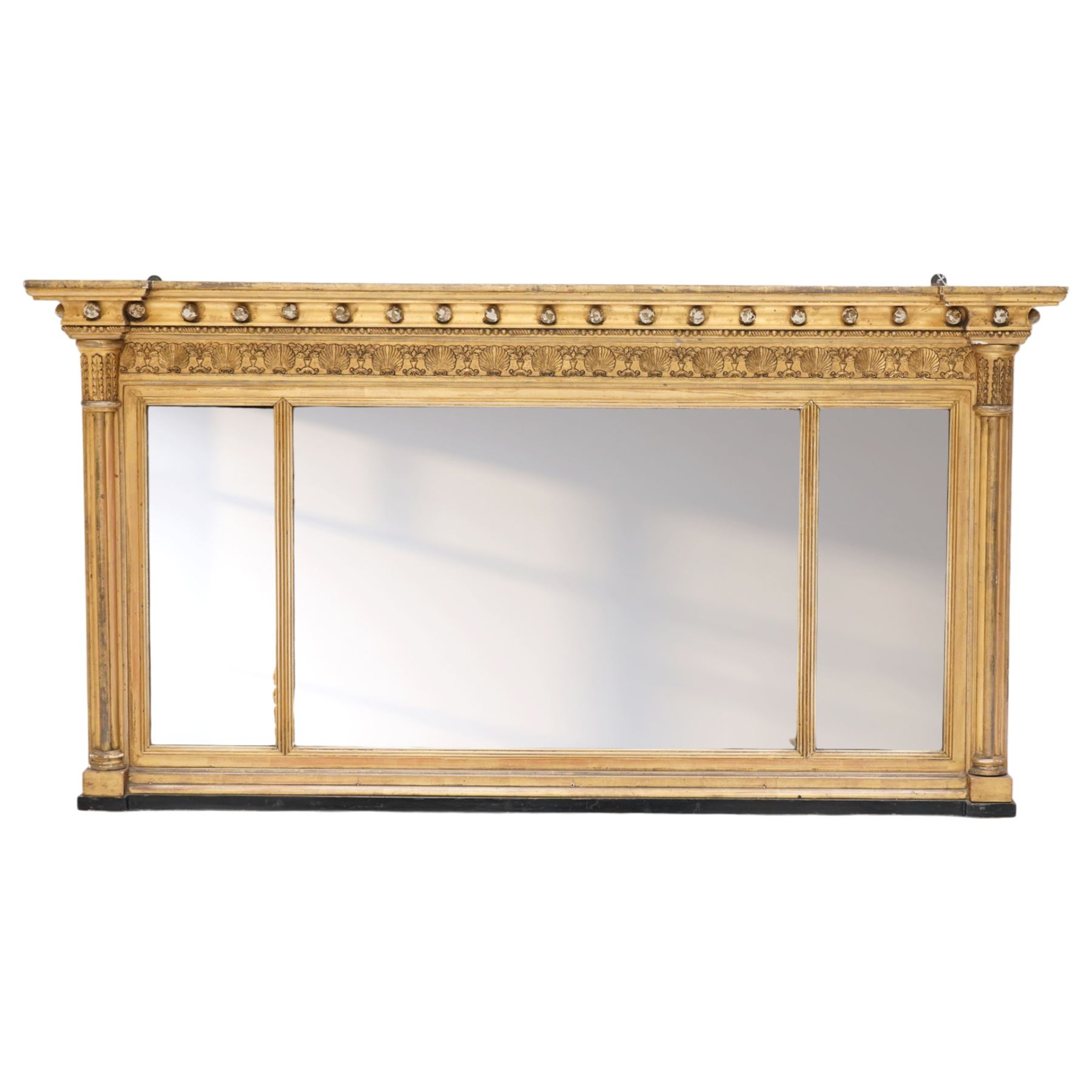 Regency period gilt and gesso framed tripartite overmantel mirror, projecting cavetto cornice applied with globular mounts, the frieze carved with repeating scallop and floral urn motifs, flanked by canted cluster-column pilasters with acanthi capitals, the triple rectangular bevelled plates within reeded slips, on skirted ebonised base
