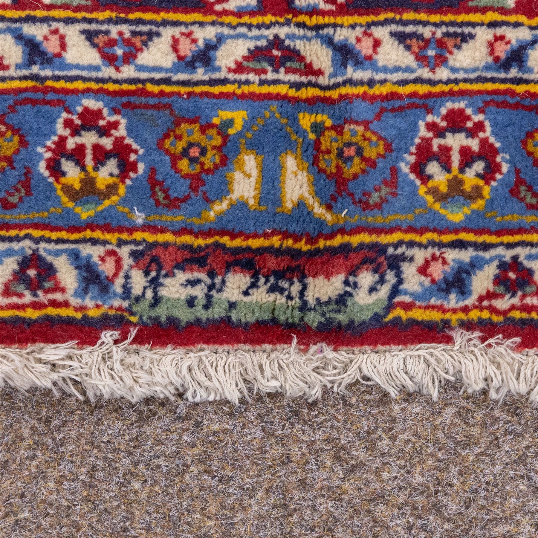 Central Persian Kashan hand-knotted carpet, madder red field with central cusped medallion in navy and ivory enclosing a floral rosette and radiating scrolling arabesques and palmettes, across a densely ornamented ground of finely drawn vinery enriched with a profusion of flowering sprays and stylized blossoms, spandrels in deep indigo and principal border of navy ground decorated with a continuous palmette and vine-scroll flanked by multiple guard stripes, with cotton fringe ends