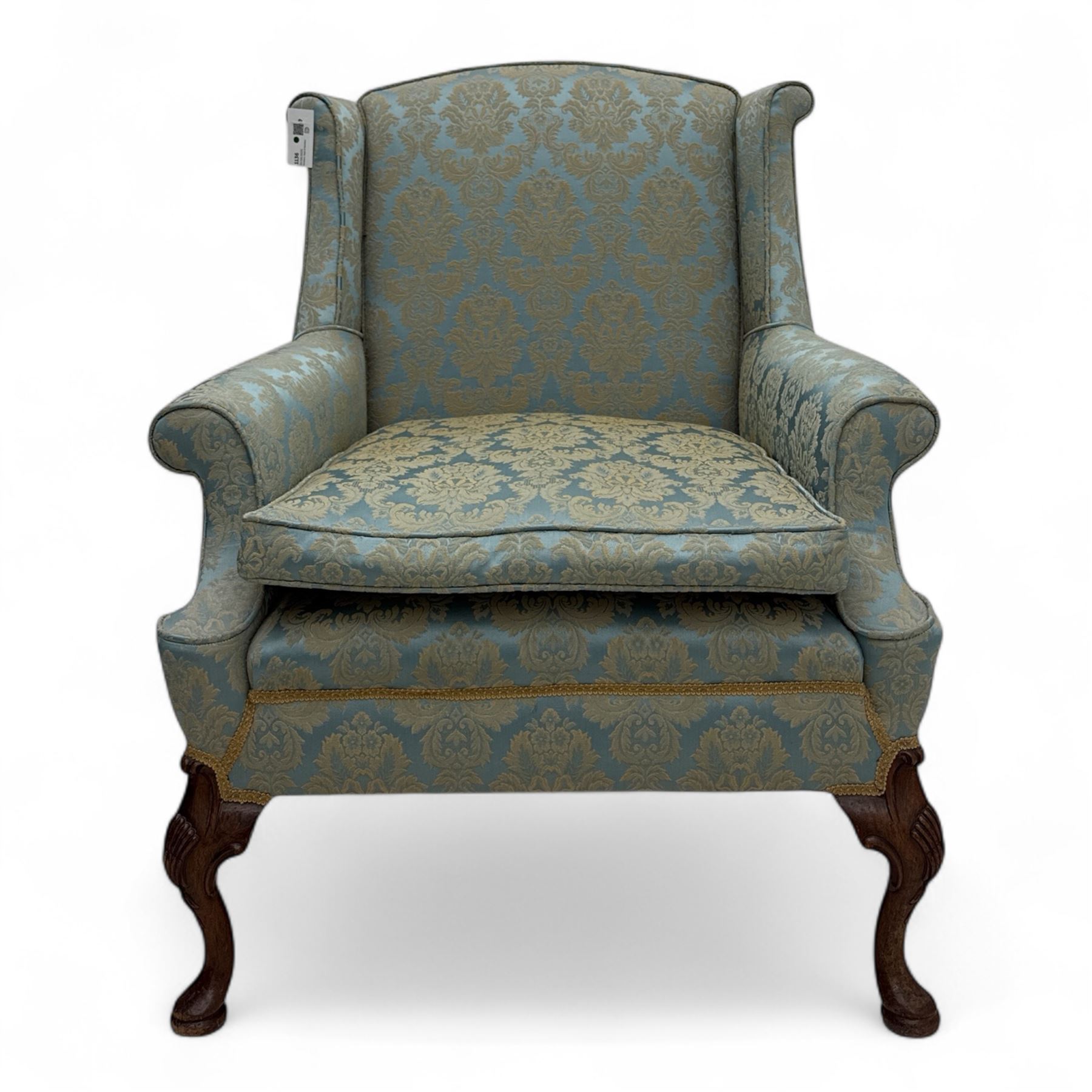 Georgian design carved walnut framed wingback armchair, upholstered in pale blue and gilt floral damask, with shaped loose seat cushion and rolled arms, raised on scallop and cartouche carved front cabriole supports terminating to pad feet