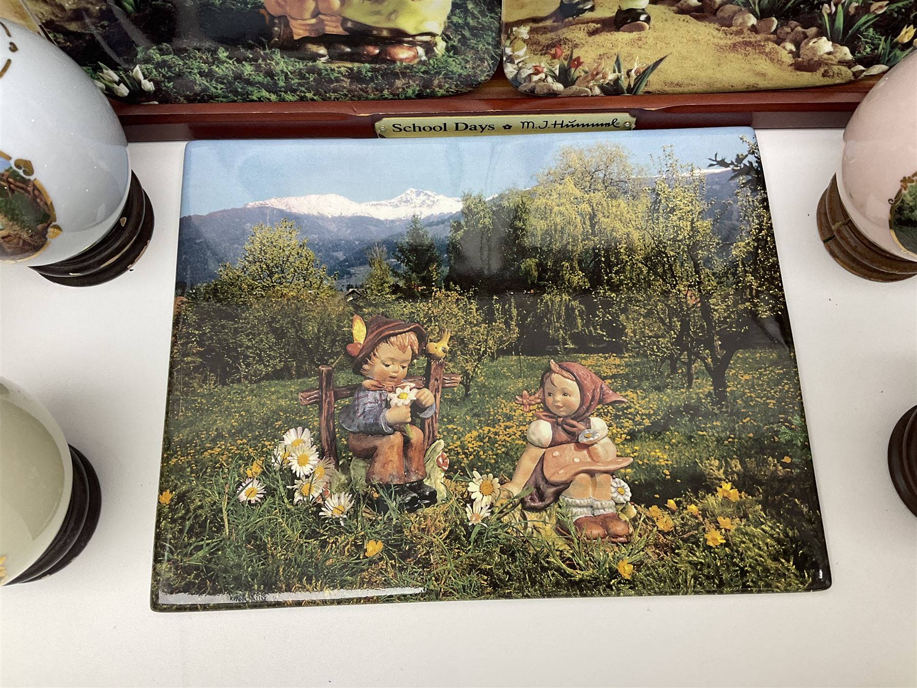 Two ceramic Hummel postcards by Goebel, both displayed on wooden easels, together nine Hummel eggs, set of four Hummel School Days plates, on wooden hanging rack, Goebel Collectors Club Member plaque etc